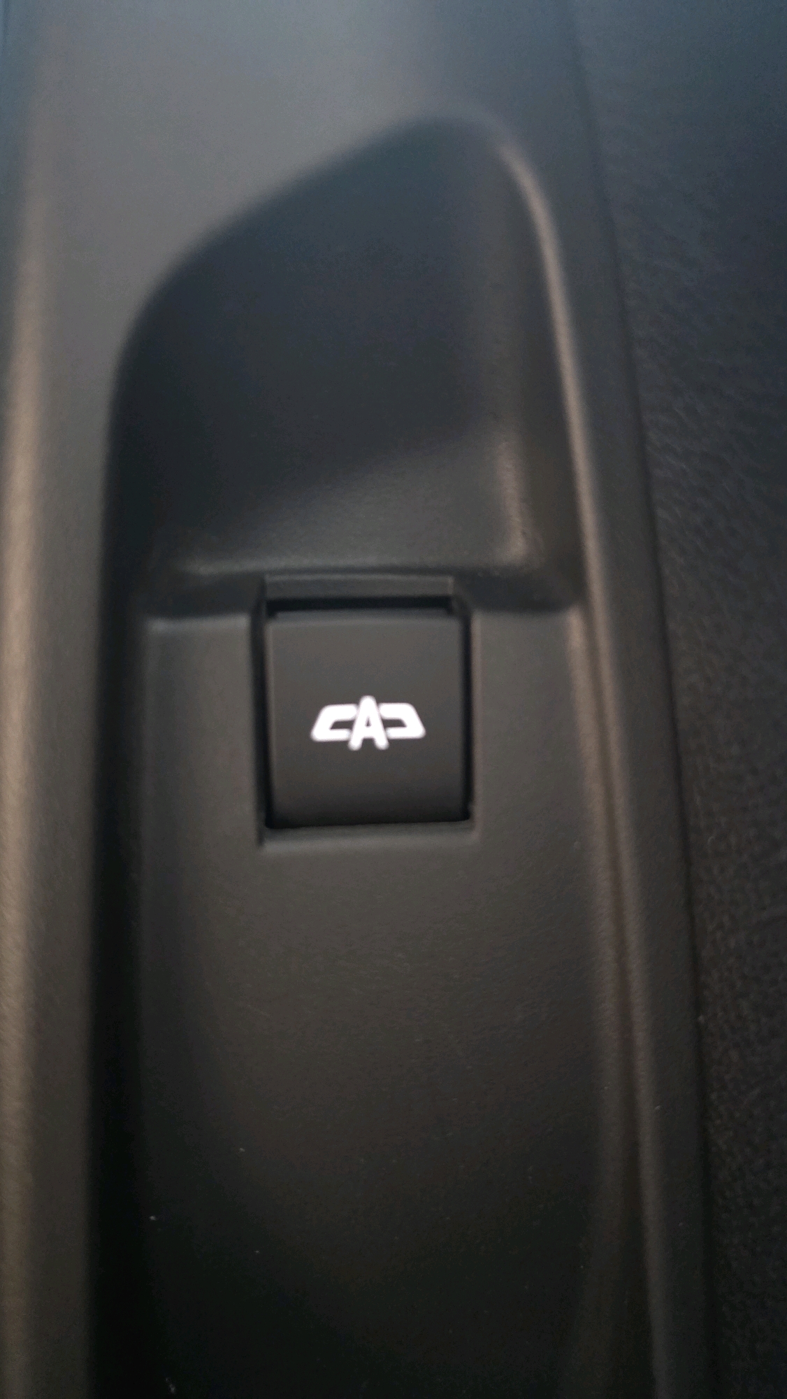 The image shows a close-up of a black button with a white symbol on it, likely inside a vehicle. The symbol appears to be a stylized representation of a convertible car's roof being lowered. The button is set within a black dashboard or interior panel. The surrounding environment is out of focus and appears to be dark and textured. The scene is not in the city of Chuy, Kyrgyzstan; it is a generic interior shot of a vehicle.