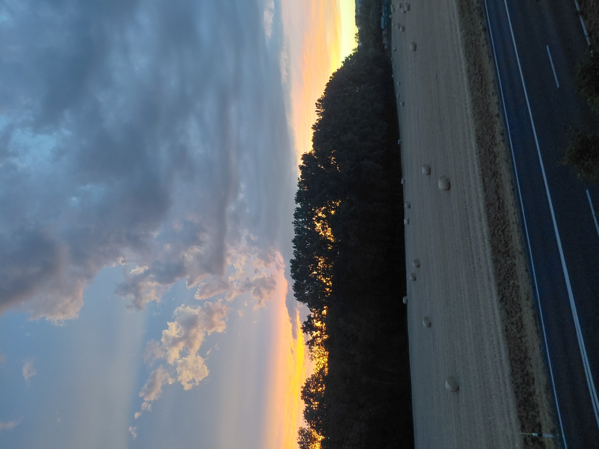 The image depicts a rural landscape scene at dusk, with a field of hay bales in the foreground. A dark treeline runs across the mid-ground, silhouetted against a vibrant sunset sky. The sky is filled with dramatic clouds, ranging from bright white and orange near the horizon to dark grey and blue overhead. A paved road with white markings runs along the bottom of the image. The hay bales are round and scattered across the harvested field. The overall impression is one of a serene, natural setting during twilight hours. There are no people or animals visible in the image. The location is identified as Ecublens, Switzerland.