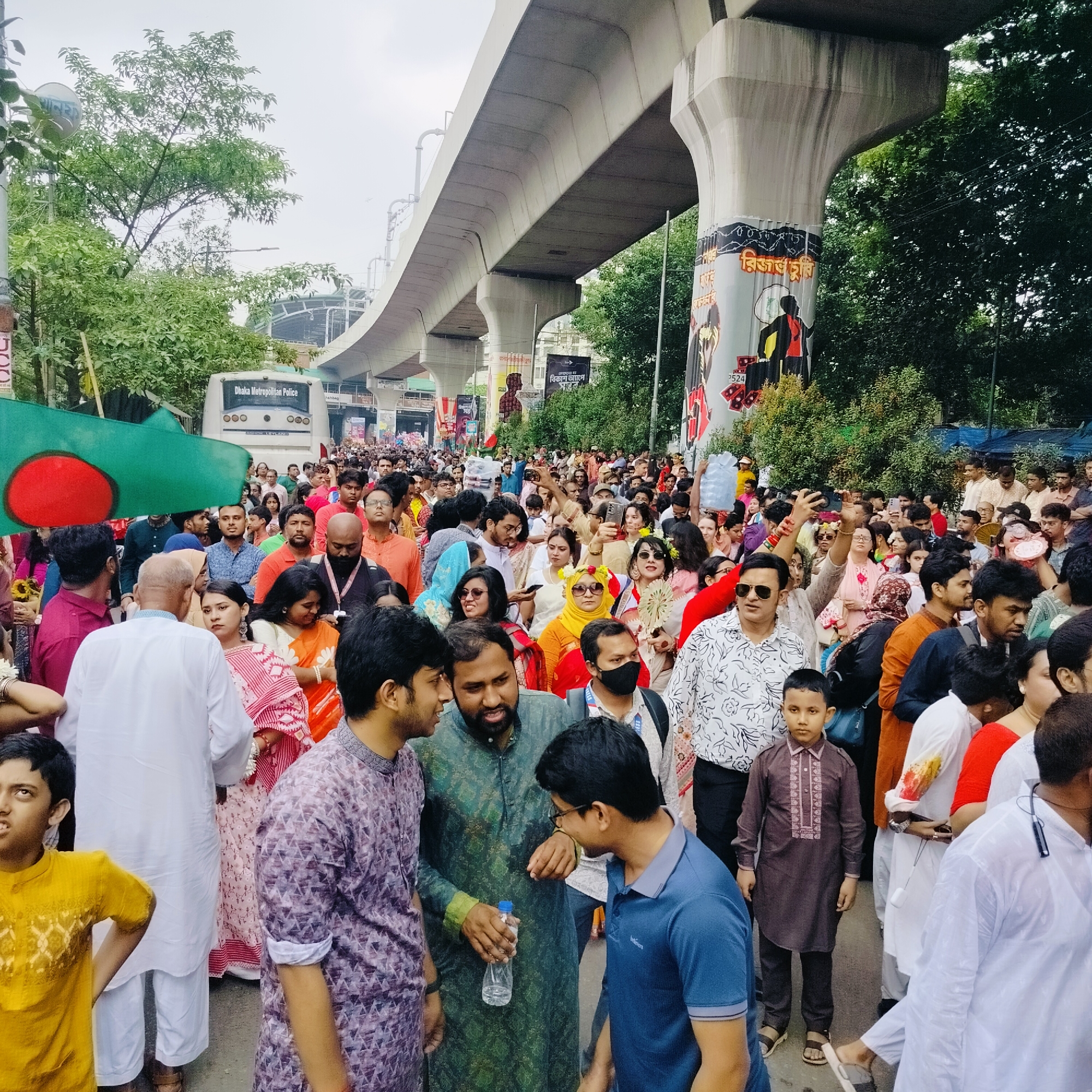 A daytime image of a dense crowd of people on a road in Dhaka, Bangladesh. The majority of people are facing forward as if they are walking together. Many people are wearing traditional clothing. To the left side, a large green and red flag of Bangladesh is being held. Above the crowd is an elevated train track. On the left, a Dhaka Metropolitan Police bus is parked. The weather is overcast.