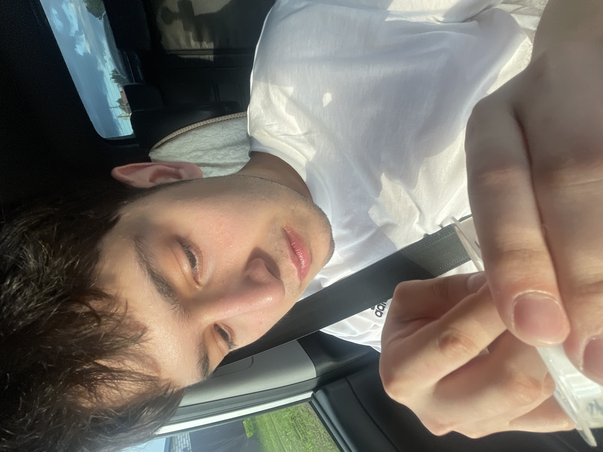 A young male with dark, messy hair and light skin is sitting in the passenger seat of a car, looking down at a small white object in his hands. He is wearing a white t-shirt with an Adidas logo. The sun is shining, casting shadows on his face and shirt. The background shows a glimpse of a suburban or rural environment through the car window, with houses and trees visible under a bright blue sky with scattered clouds. The interior of the car is dark, with a visible headrest and part of a seatbelt. The image appears to be taken outdoors, during the daytime, possibly in the city of Gavrilovka, Russia, based on the provided context.