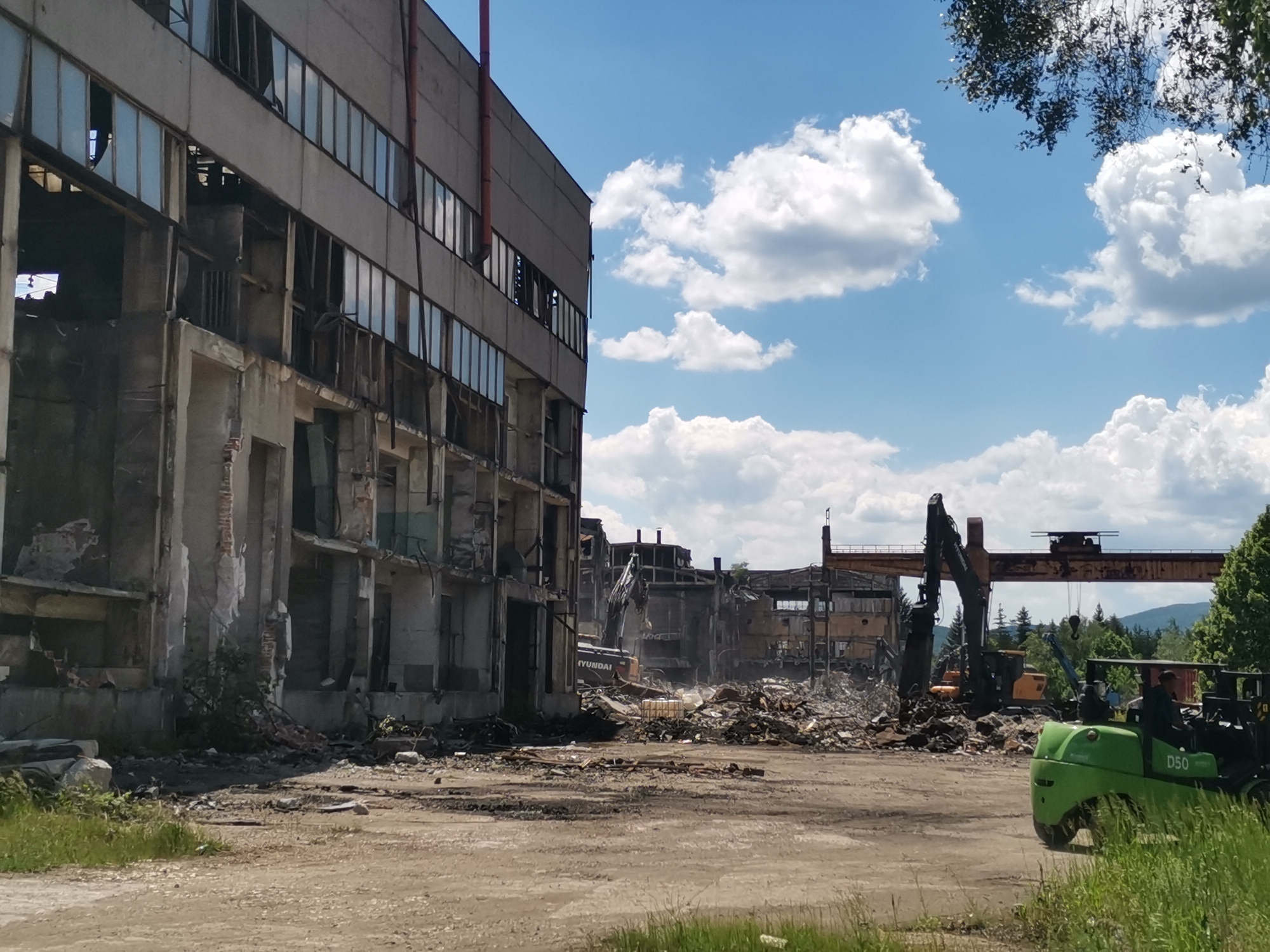The image depicts a demolition site in Zhivkovo, Bulgaria. The skeletal remains of a large, multi-story building stand on the left, partially gutted with exposed interiors and broken windows. Debris is scattered across the foreground. Heavy machinery, including excavators and a large crane, are amidst the wreckage. A green forklift is positioned on the right side of the image. The sky is blue with scattered white clouds. Trees border the right side of the frame and appear in the background.