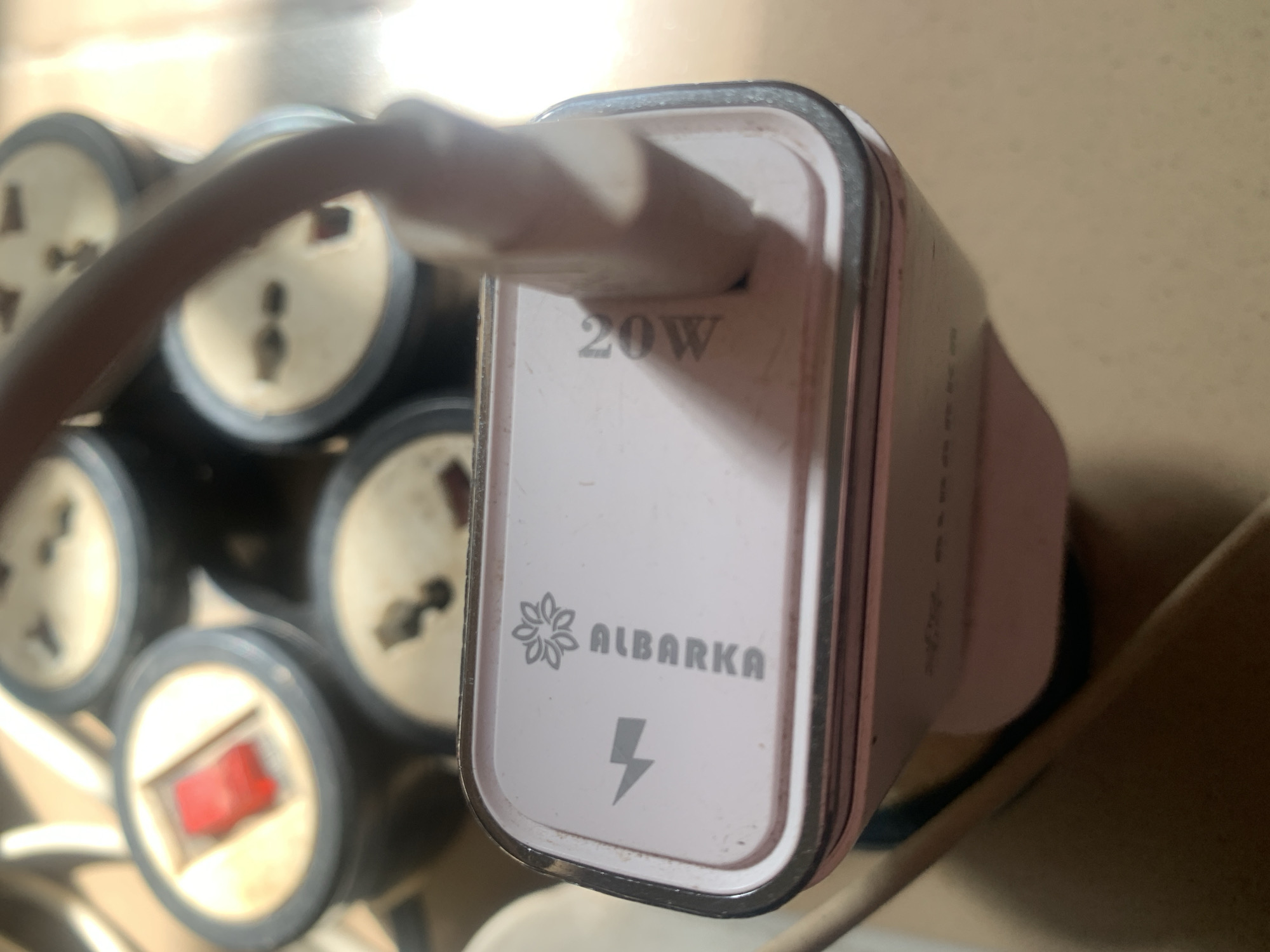 The image shows a close-up of a white and pink power adapter plugged into a multi-socket electrical extension. The adapter has the branding "ALBARKA" and a "20W" designation. In the background, several electrical sockets with red switches are visible, suggesting the adapter is in use in an indoor setting. The overall scene appears to be an electrical setup for charging devices. There is no information about Jauro Yinu, Nigeria in the image.
