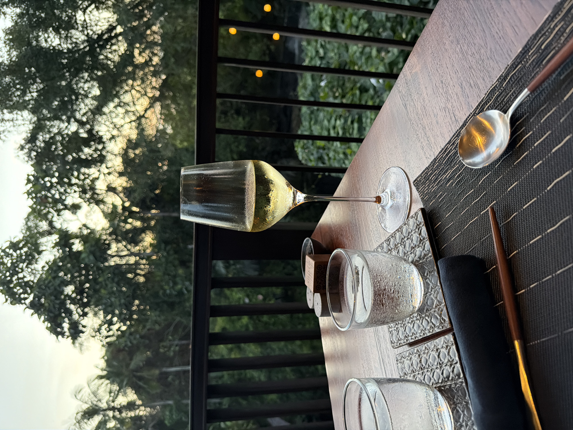 The image shows a table setting on what appears to be a balcony or open-air dining area. The scene includes a partially filled wine glass, several empty drinking glasses, a spoon, a napkin, and chopsticks, all placed on a patterned placemat on a wooden table. Outside the balcony, there is lush greenery. The lighting suggests either dusk or dawn. Without additional information, it's not possible to verify the precise location within Kediri, Indonesia.