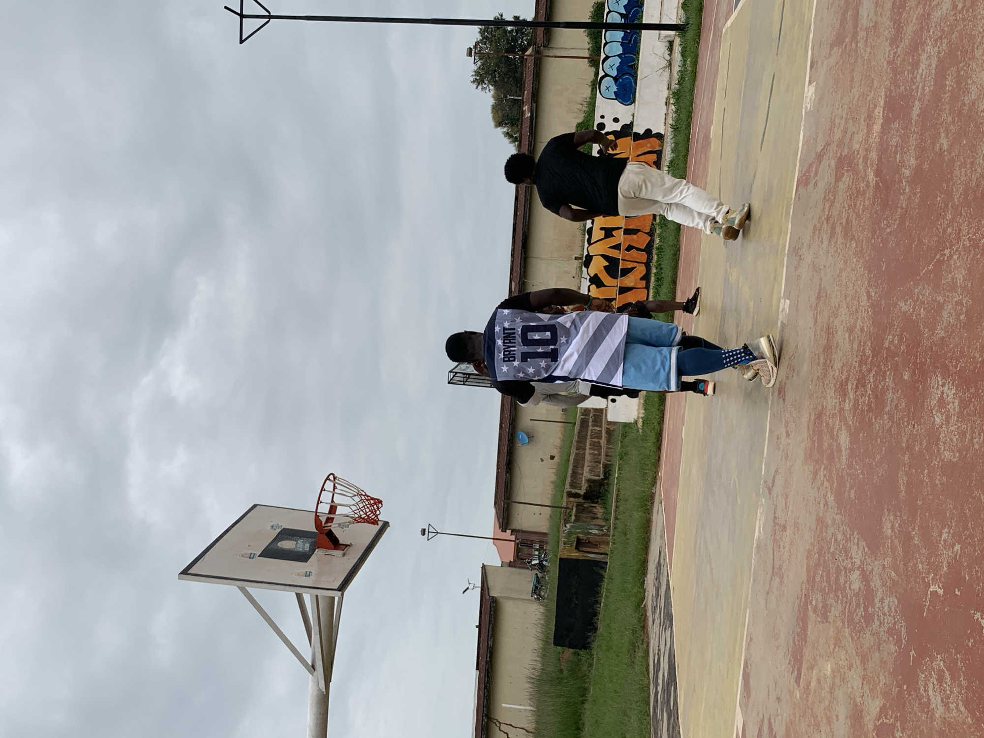 This image captures an outdoor basketball court in Gura Top, Nigeria, under a cloudy sky. In the foreground, three individuals are engaged in a basketball game. The person closest to the viewer is wearing a black shirt with white stars and stripes and the number "10" on the back, along with blue basketball shorts and gold sneakers. Another person, to the right, wears a black t-shirt and light-colored pants, also in sneakers. A third person, partially visible between them, is in motion. The court itself is weathered, with sections of red and yellow concrete, and white lines marking the boundaries.
In the background, a basketball hoop with a red net is mounted on a white backboard. Beyond the court, a low-slung building with a reddish-brown roof is visible, showing signs of age and wear, including visible cracks. Adjacent to the building, there is a patch of green grass and some steps leading up. Graffiti with orange and blue lettering adorns a low wall behind the court. A tall, thin pole with a triangular structure at the top stands near the right edge of the frame, suggesting it might be a lighting pole. The overall scene depicts a casual game of basketball in a somewhat rundown but functional outdoor court.