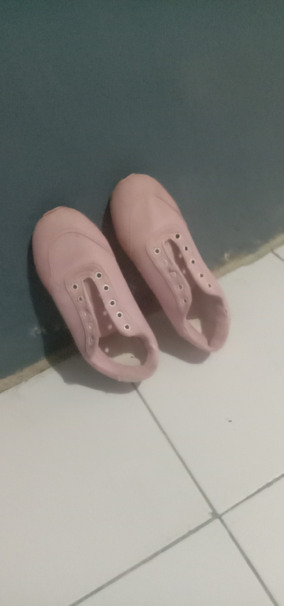 Two pink, canvas slip-on shoes without laces are lying on a white tiled floor next to a dark teal wall. The shoes are positioned in the bottom half of the frame, with the left shoe closer to the viewer and the right shoe slightly behind and to the right of it. Both shoes are empty and their laces are absent, as indicated by the visible eyelets. The floor tiles are arranged in a grid pattern, with visible grout lines. The wall behind the shoes appears to be a solid, dark color with a matte finish. The lighting in the scene is dim, casting shadows from the shoes onto the floor. This scene appears to be an interior setting, possibly a room or hallway in a residential or commercial building. There is no indication of the specific location within Kendari, Indonesia.