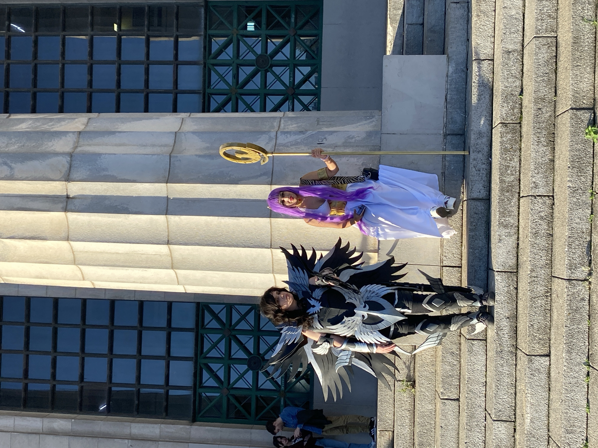 Two individuals in cosplay attire are posing on stone steps adjacent to a building with large windows in Buenos Aires, Argentina. One individual, located lower on the steps, wears a dark costume with large wing-like extensions. The second individual, positioned above the first, is clad in a white gown and purple wig and holds a gold-colored staff. In the background, a building with large, grid-patterned windows and stone facade is visible. Other people can be partially seen at the bottom of the image. The location is most likely near the National Library.