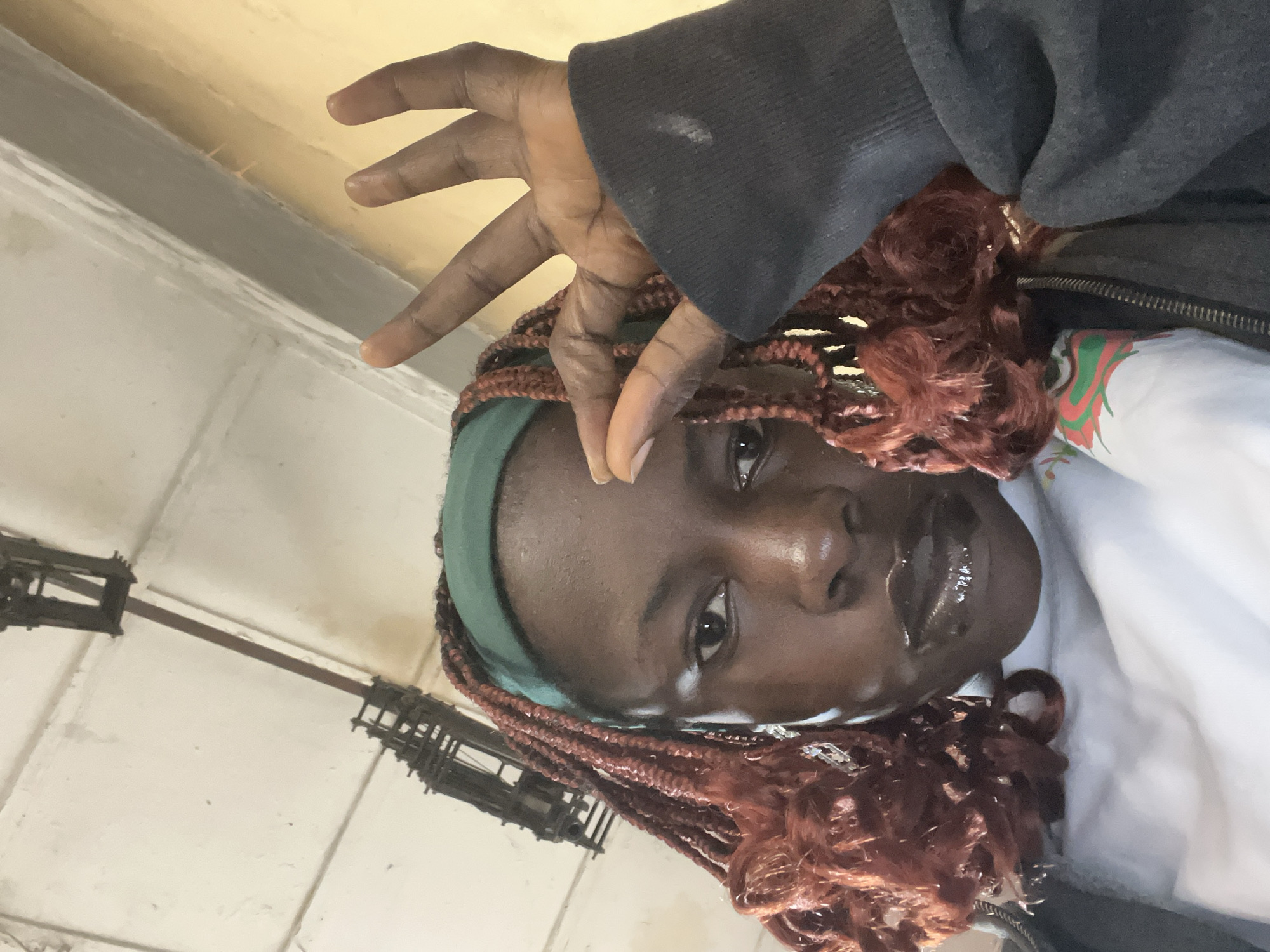 A close-up, low-angle, selfie-style shot captures a young Black woman from the shoulders up, looking directly at the camera. She is wearing a dark grey hooded jacket over a white t-shirt with a colorful graphic print on the left side. A green headband adorns her head, and her dark brown braided hair with reddish highlights is visible on either side of her face. She is wearing dark, glossy lipstick. Her right hand is raised, with her thumb and index finger forming a circle, while her other fingers are extended upwards. Her left hand is partially visible near her forehead.
The background suggests an indoor setting with a light-colored ceiling featuring industrial lighting fixtures and exposed pipes. The walls are painted in a pale yellow hue. The overall lighting is somewhat dim, with a warm cast. The composition is framed by the woman's head and shoulders, with the background blurred.