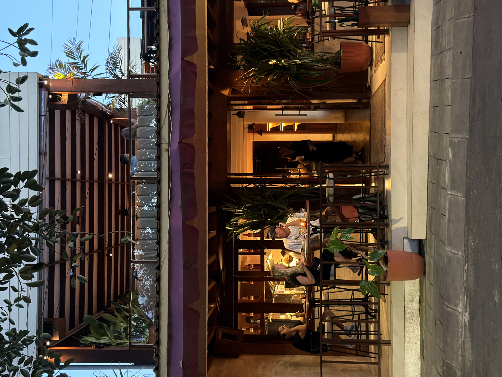 The image shows an outdoor seating area of a restaurant in North Kuta, Indonesia, at dusk. People are seated at tables, dining and conversing. The restaurant's facade incorporates wooden beams, striped awnings, potted plants, and glass elements. The street is paved with stone blocks. The lighting is warm, highlighting the interior space.
