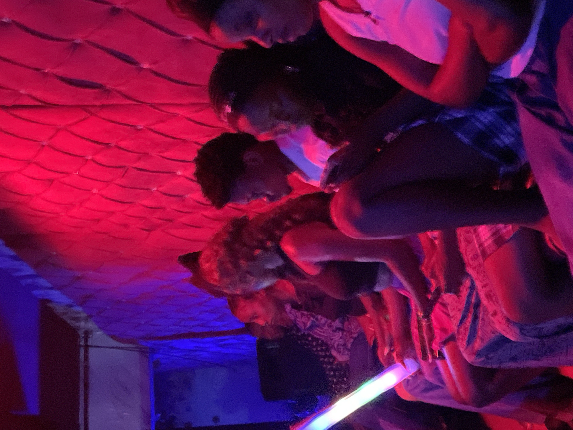 A group of young people are seated in a dimly lit indoor space, likely a club or lounge, in Durumi, Nigeria. The environment is characterized by intense red and blue lighting, creating a vibrant and somewhat diffused atmosphere. A large, textured wall paneling, illuminated by the red light, dominates the background.
In the foreground, several individuals are visible, all appearing to be of African descent. They are seated closely together, suggesting a social gathering. One person on the far left is holding a long, cylindrical object that emits a colorful glow, possibly a light stick or a novelty item. The individuals are dressed casually; one person wears a black tank top, another a white shirt. Some are looking at their phones, indicating a relaxed, contemporary social interaction. The overall impression is one of a lively, youthful evening outing in a Nigerian urban setting.