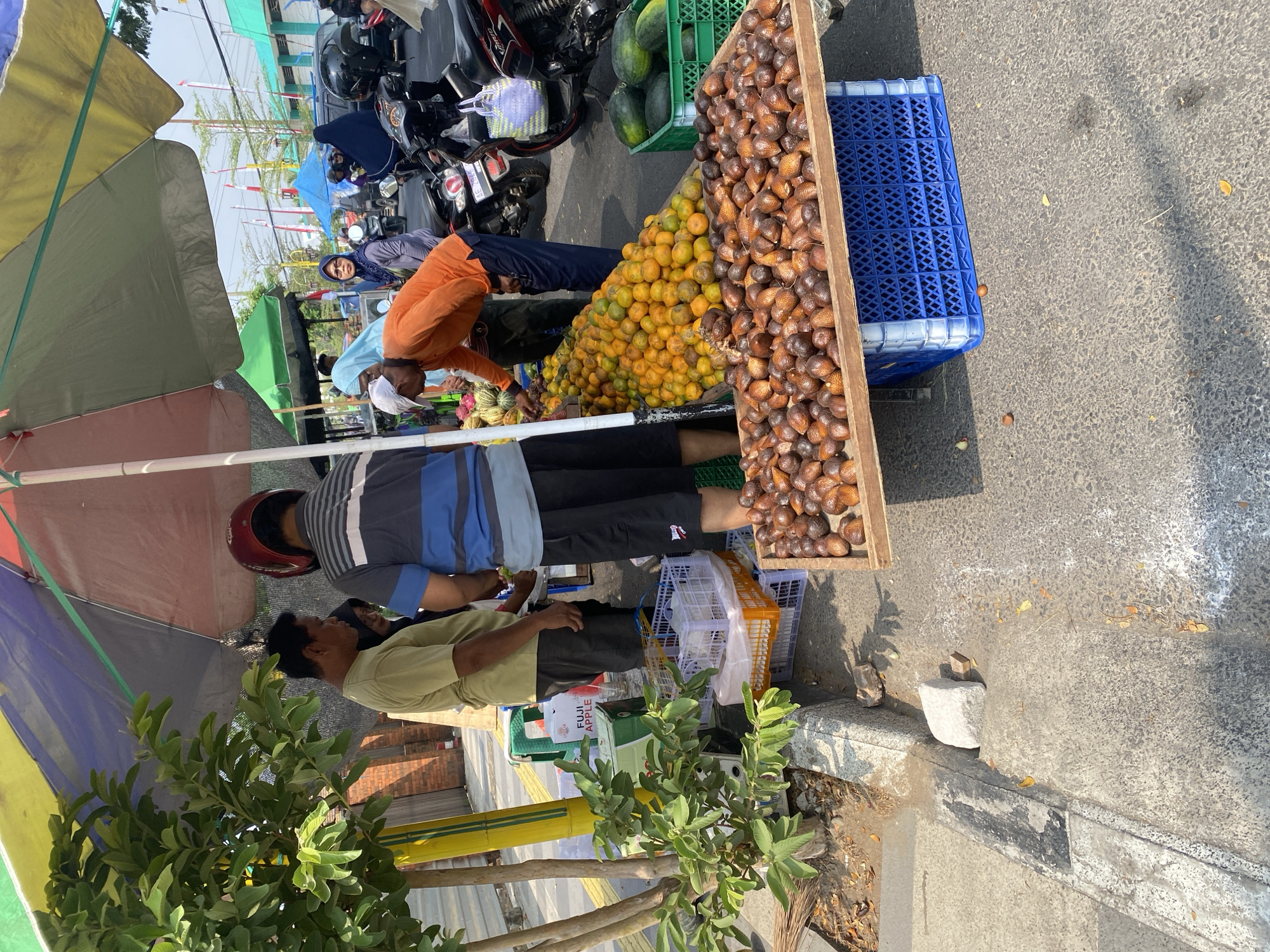 A street vendor in Sempol, Indonesia, displays fruit for sale under a colorful umbrella. A pile of oranges sits in front of a wooden plank topped with salak fruit. Behind them, a man in an orange shirt reaches for fruit. In the background, motorcycles and cars are parked along the road. Green crates of watermelons are visible to the right. A small tree with green leaves stands to the left, next to a yellow pole. The ground is paved with concrete, and dry leaves are scattered on the surface.