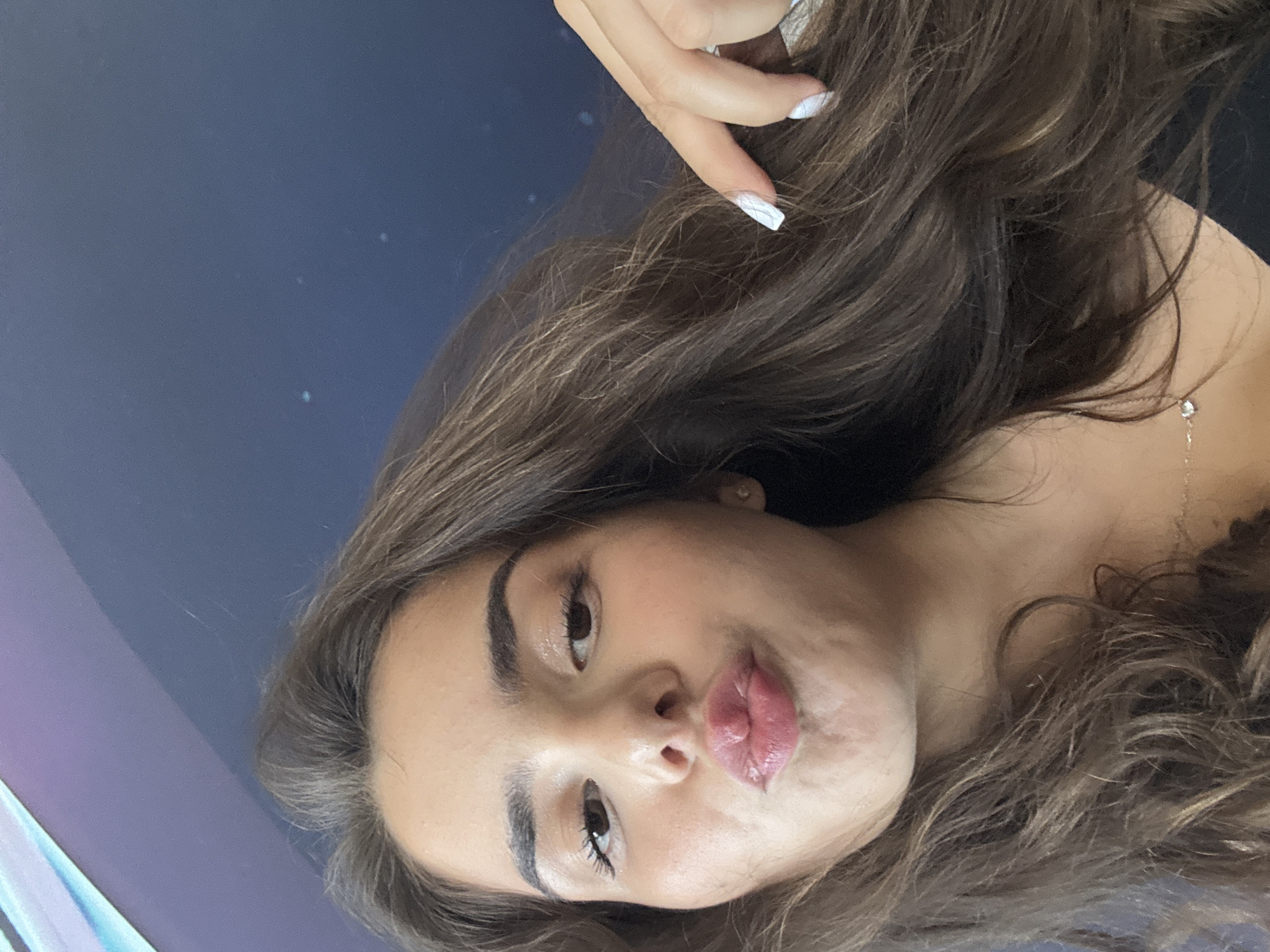 The image is a close-up selfie of a young woman with long, brown hair. She is making a kissing face, with her lips pursed. Her eyes are dark brown and her eyebrows are well-defined. She is wearing light makeup, including mascara and lip gloss. A hand with white nail polish is visible in the upper right corner of the image, touching her hair. The background is a dark blue wall, possibly with some out-of-focus lights. The setting does not provide any specific information about the city of Shumen, Bulgaria, or any discernible landmarks. The image captures a moment of personal expression from the subject.