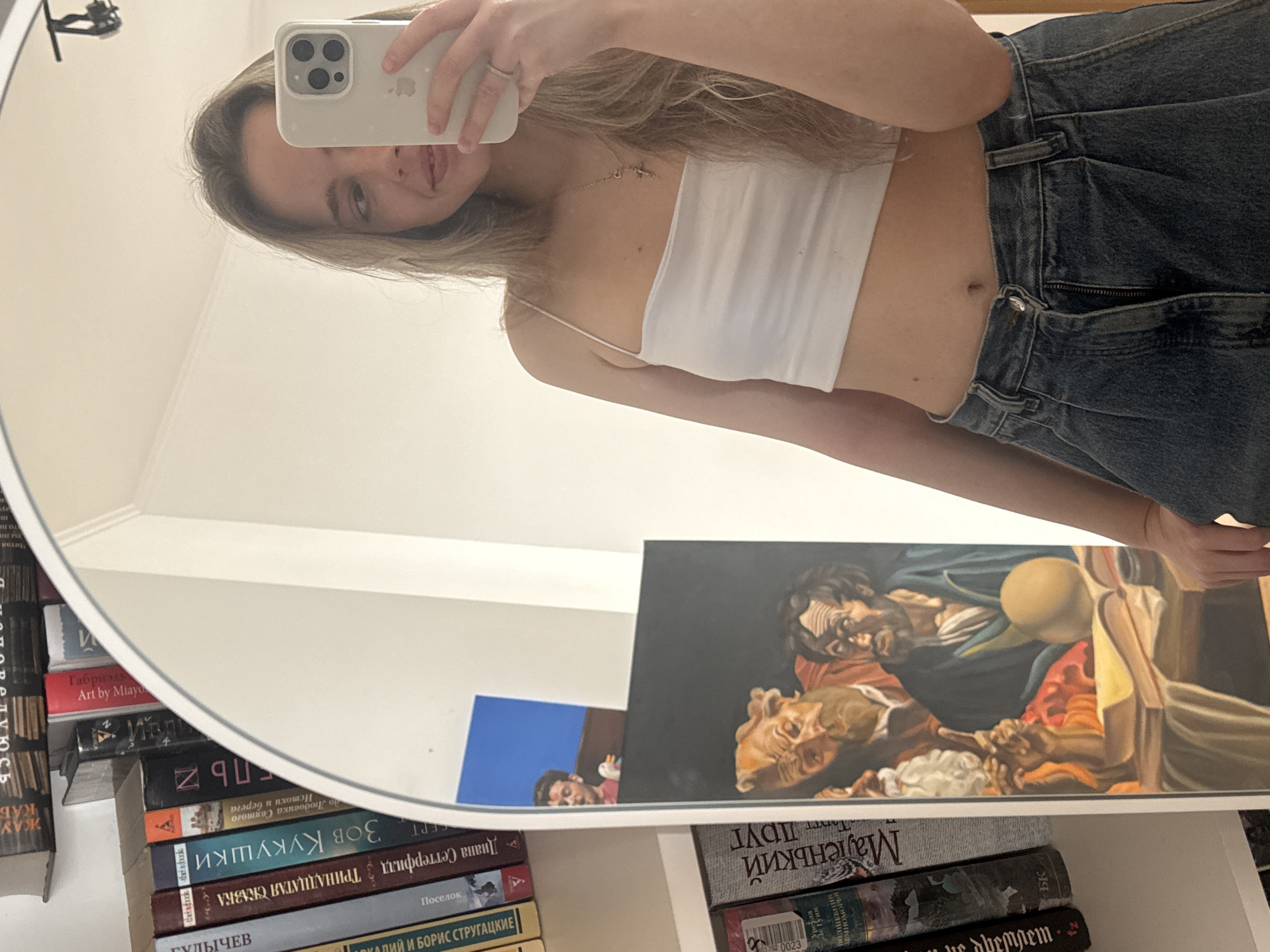 The image is a selfie taken in a mirror. A young woman with long blonde hair is holding an iPhone to capture the reflection. She is wearing a white crop top and blue jeans. Her midriff is visible. The mirror reflects part of a room, specifically a shelf containing books and artwork. The books are stacked with their spines facing the mirror. Titles in Cyrillic script are visible, including "Тринадцатая сказка" and "Маленький Друг". Several paintings are visible, including a large piece depicting several men. The location of Khimki, Russia is not explicitly confirmed in this image.