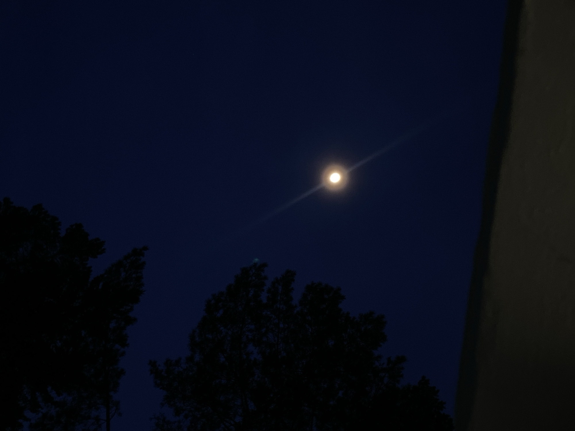 This image depicts a night scene in Chaikovsky, Russia, specifically focusing on the sky. The primary subject is a bright, circular, yellowish-white celestial body, consistent with the moon, positioned in the upper-middle left quadrant of the dark blue-black sky. A distinct, diagonal lens flare extends through this celestial object. In the lower right section of the frame, dark silhouettes of dense tree foliage are visible, indicating the presence of trees, possibly conifers, against the night sky. A dark, horizontal band occupies the top portion of the image, representing an overhead obstruction such as a roofline or architectural element, partially obscuring the upper sky. No people, specific actions, or interactions are visible. The precise location within Chaikovsky is not discernible from the visual information provided.