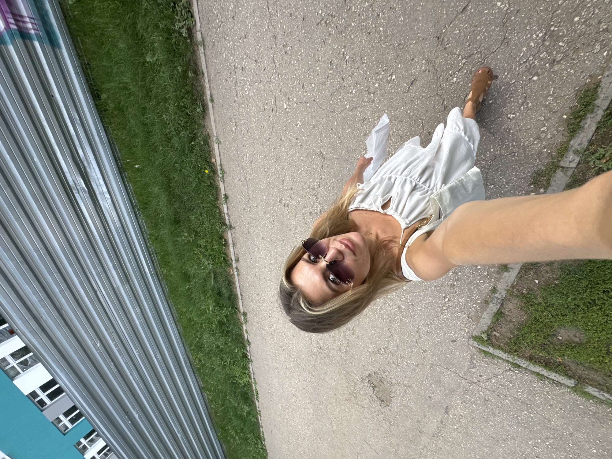 A young woman with blonde hair and sunglasses is taking a selfie outdoors. She is wearing a white dress and is holding a white bag in her right hand. She is standing on a paved surface next to a grassy area. In the background, there is a corrugated metal fence and a building with windows. The scene appears to be a city street or sidewalk.