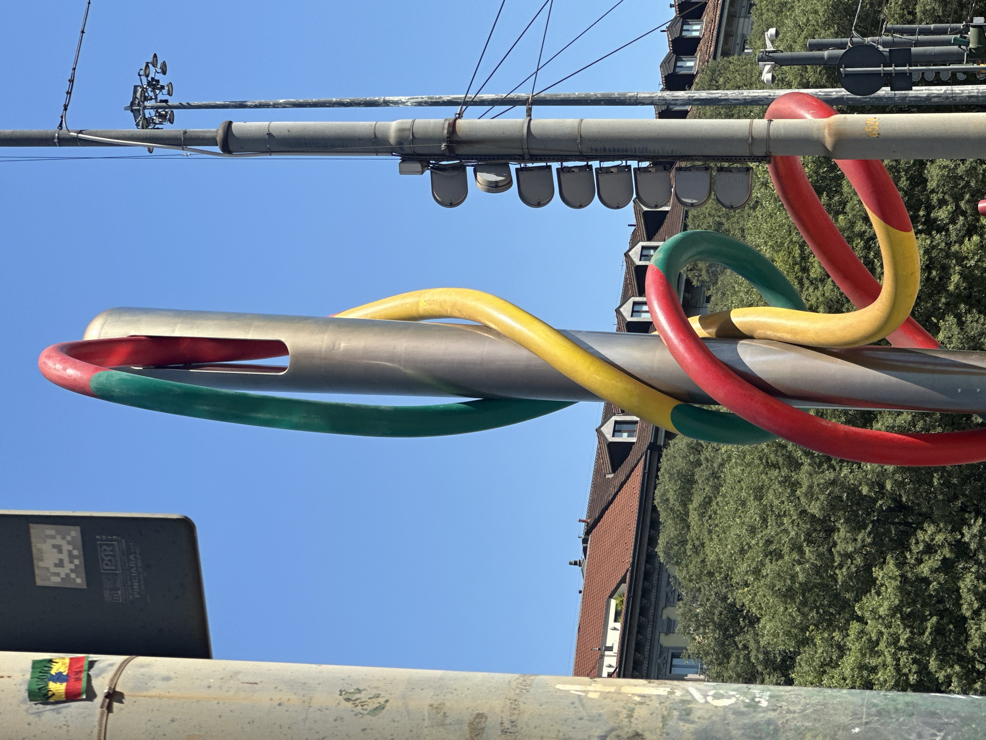 The image depicts a giant needle and thread sculpture set against a blue sky and a backdrop of buildings and trees. The needle is metallic and silver in color, with a large eye containing three loops of colored thread: green, yellow, and red. The sculpture is mounted on metal posts, with streetlights and utility lines visible above. The sculpture is located at Piazzale Cadorna in Milan, Italy, and is a well-known landmark in the city.