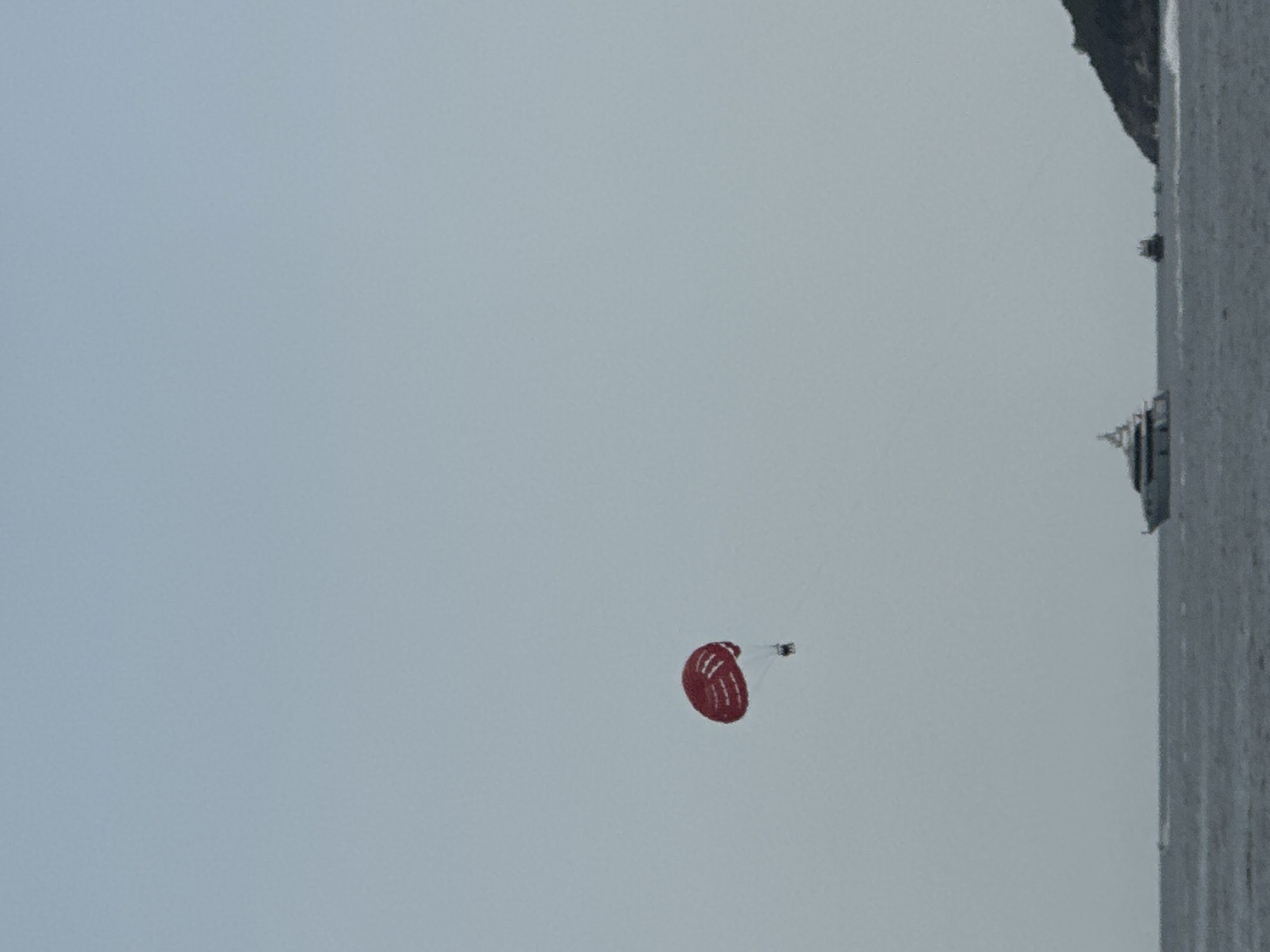 A parasail with a red canopy and white stripes is visible against a pale sky. Below the parasail, a person is suspended, appearing to be towed. In the distance, on a calm, gray body of water, a large white yacht with multiple decks sails to the left. Further to the right, a smaller boat is visible, and beyond that, a dark green, hilly landmass juts into the frame from the right side of the image, suggesting a coastal location. The overall scene depicts a recreational activity on the water, likely near a coastline.