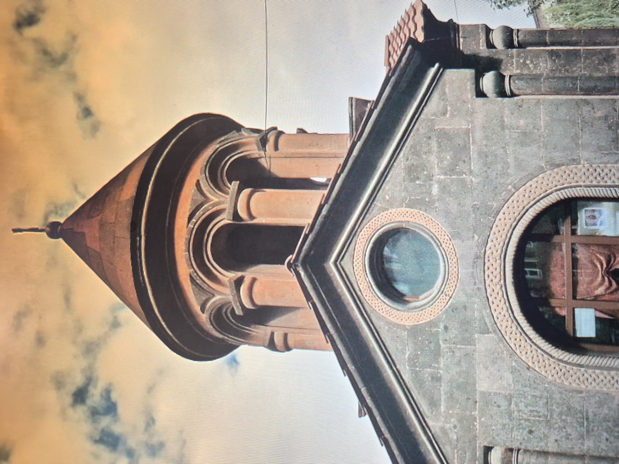 The image features a close-up view of the Holy Mother of God Church in Yerevan, Armenia. The architecture is distinct, showing a conical spire with decorative archways. The building is constructed from stone, with circular windows framed with intricate detailing. A portion of the roof is visible, showcasing dark-colored stone elements. The sky behind the structure is light, with hints of clouds.