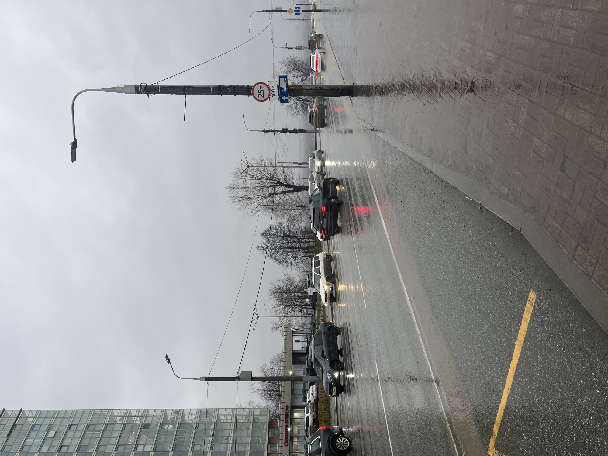 The image depicts a street scene in Perm, Russia on a rainy day. A line of cars is traveling on a wet asphalt road. A pedestrian walkway made of grey rectangular tiles is adjacent to the road. Streetlights are positioned along the road. Traffic signs are visible on the utility poles. Several buildings and bare trees are also in the background. The sky is overcast. The overall environment suggests an urban setting.