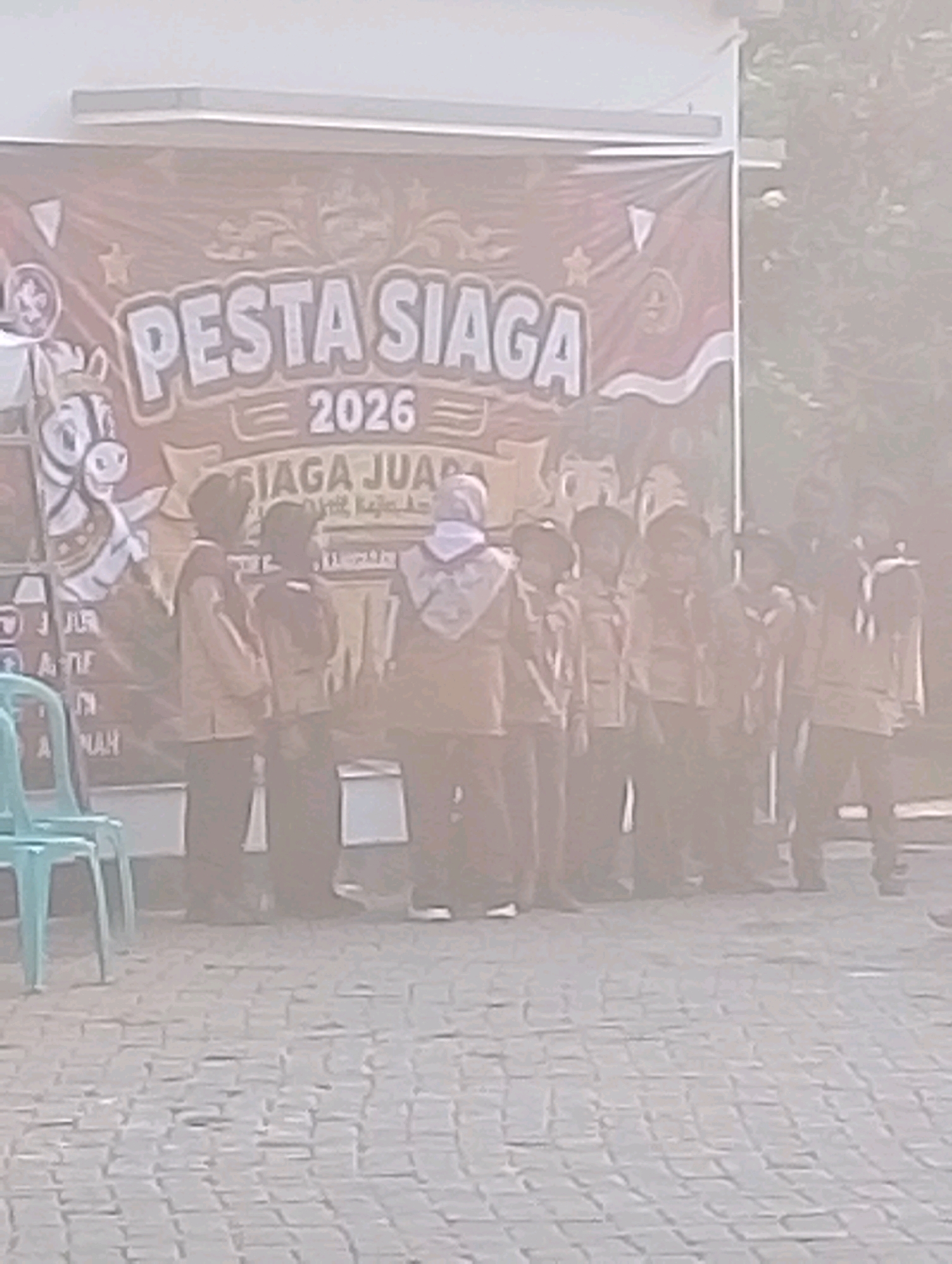 A group of children in uniform stand in front of a banner that reads "PESTA SIAGA 2026". The banner also features decorative elements and a cartoon image of a horse. The children appear to be participating in an event, possibly a scouting or youth gathering. In the background, there are trees and some architectural elements. The ground is paved with interlocking bricks. The scene takes place outdoors, possibly in a public space or schoolyard in Probolinggo, Indonesia.