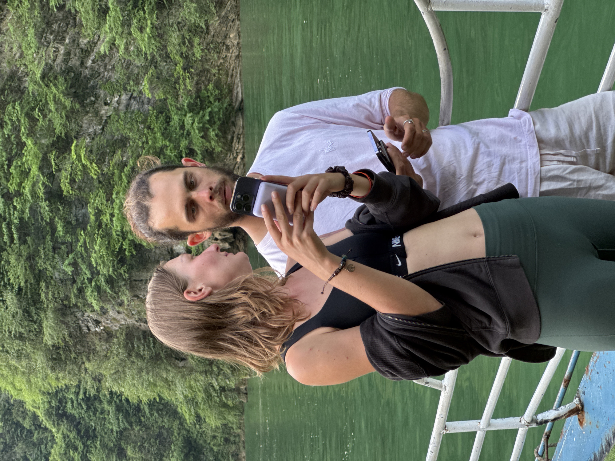 A man and a woman are taking a selfie on a boat on a body of water. The woman is holding a phone and pointing it towards herself and the man. The man is holding a second phone. They are both looking at the phone in the woman's hand. They are on a boat on the emerald green waters of the Nho Quế River, adjacent to Đồng Văn Commune, Vietnam. The surrounding environment consists of a cliff wall covered in lush, verdant vegetation. The boat has a painted blue and white railing.