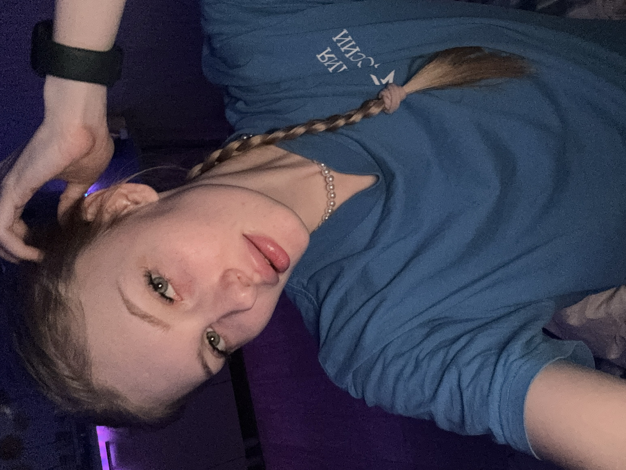 The image, taken in Кондратово, Russia, depicts a young woman lying down and capturing a selfie. She has light-colored hair styled in a long braid that extends over her shoulder, secured with a pink or light-colored hair tie. Her eyes are green or hazel, and she maintains a direct gaze at the camera. Her lips are slightly parted, showcasing a subtle pink tint.
She is wearing a blue long-sleeved t-shirt featuring Cyrillic text on the front, which appears upside down and mirrored, reading "ИИХОССИИ БИУ". A beaded necklace is visible around her neck, and a black band or bracelet adorns her left wrist.
The setting is indoors, with low ambient lighting, suggesting evening or nighttime. The background is predominantly dark with purplish hues, and a bright purple light source is visible on the left side of the frame. The woman's expression is neutral to slightly pouting, conveying a relaxed demeanor.