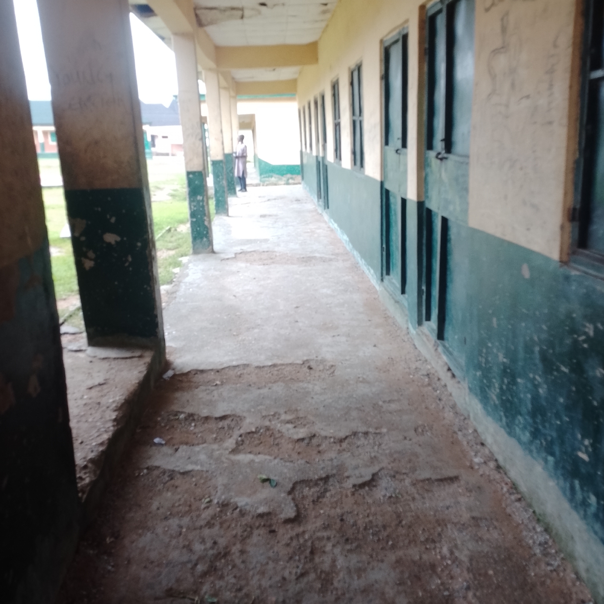 The image shows a weathered, outdoor hallway in what appears to be an institutional building, possibly a school or clinic, in Serti, Nigeria. The hallway has a concrete floor that is cracked and worn, with patches of exposed dirt and debris. To the right, a row of identical doors with windows is visible along the wall, painted a dark green with a lighter green stripe below the windows. The wall above the green stripe is a light yellow color. To the left, a thick concrete pillar is prominent, with faded writing on its surface. Beyond the pillar, glimpses of green grass and other buildings can be seen. A single person, wearing a light-colored robe and dark trousers, stands further down the hallway on the left side, facing away from the camera. The overall impression is one of disrepair and neglect.