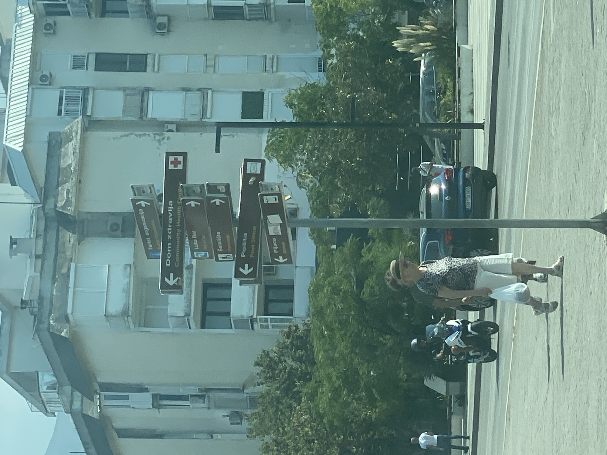 In the city of Bar, Montenegro, a directional signpost stands prominently on a street corner. The signpost displays multiple signs pointing in various directions, indicating destinations such as "Turist organizacija" (Tourist Organization), "Dom zdravlja" (Health Center) with a red cross symbol, "Luka Bar" (Bar Port) with a symbol of a ship, "Setalište" (Promenade), "Pošta" (Post Office), and "Pijaca" (Market). Behind the signpost, a modern apartment building with multiple balconies and windows is visible. On the street in front of the building, a woman wearing a patterned shirt and white shorts walks with a plastic bag in her hand. A person on a motorcycle is seen in the background, and other vehicles are present on the road. Lush green trees and tall grass plants frame the scene. The overall environment suggests a sunny day in an urban setting.