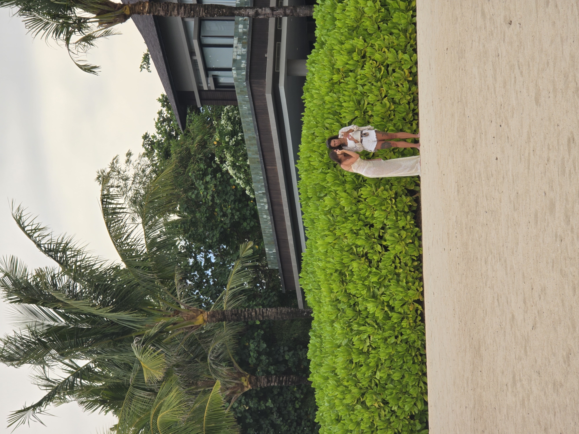 The image, taken in Nusa Dua, Indonesia, shows two women in a lush outdoor setting. One woman, wearing a full-length white gown, stands next to a second woman who is wearing a white jacket and shorts outfit. They are positioned in front of a dense green hedge. In the background, a modern building with windows and a dark-colored facade is visible. The building is partially obscured by trees and palm trees. A sandy area is visible in the lower right corner. The sky is overcast.