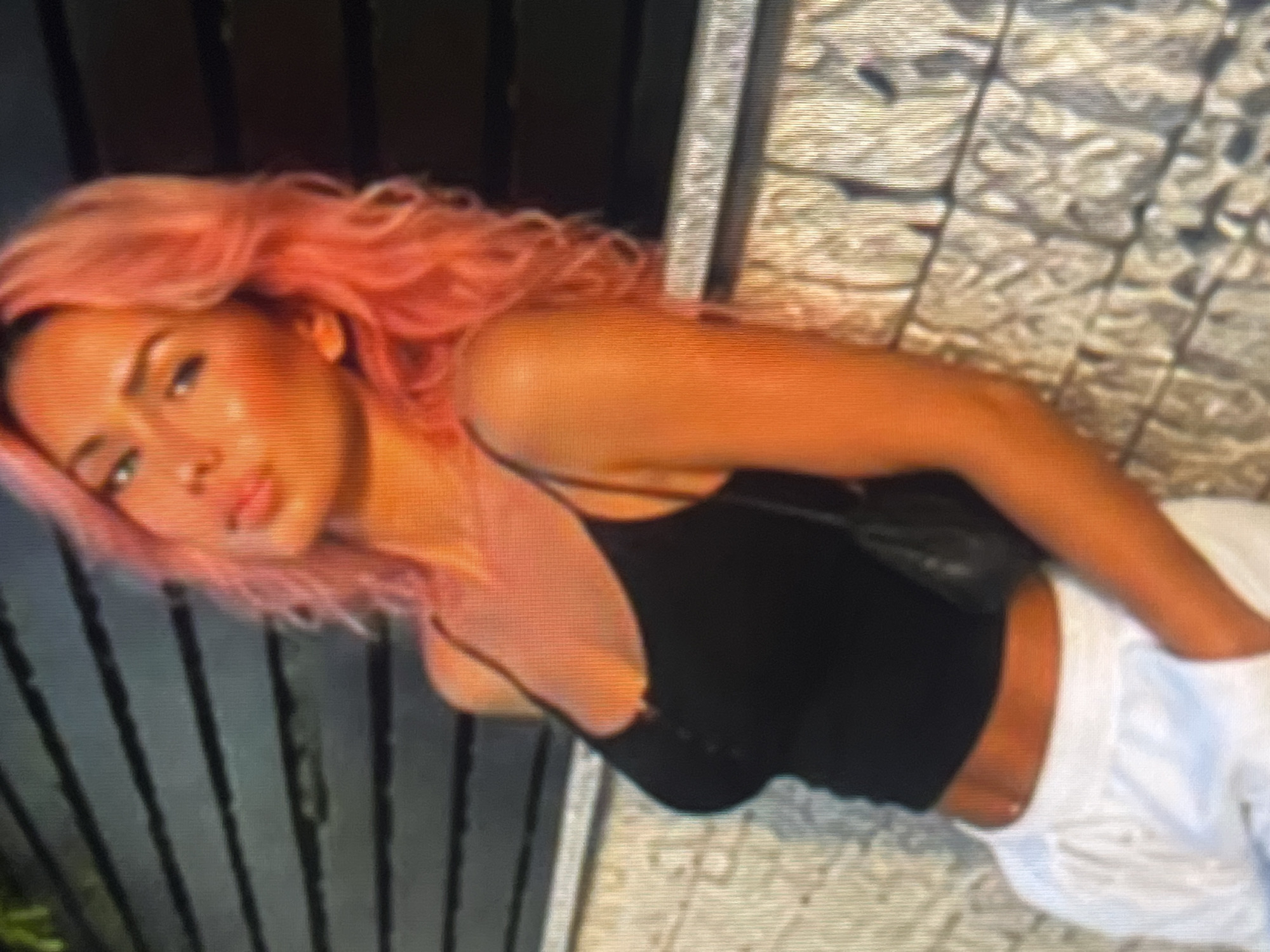 A young woman with vibrant pink hair, wearing a black tank top and white pants, leans against a textured wall in an urban outdoor setting. She is positioned on the right side of the frame, with her body angled towards the viewer. Her gaze is directed upwards, and her lips are slightly parted. The background consists of horizontal slats of a dark material, possibly a fence or wall, and the textured surface she is leaning on appears to be made of rough, patterned stone or concrete. A small black purse hangs from her shoulder. The lighting suggests it is either sunrise or sunset, casting a warm glow on the scene. The location is an unknown urban environment.