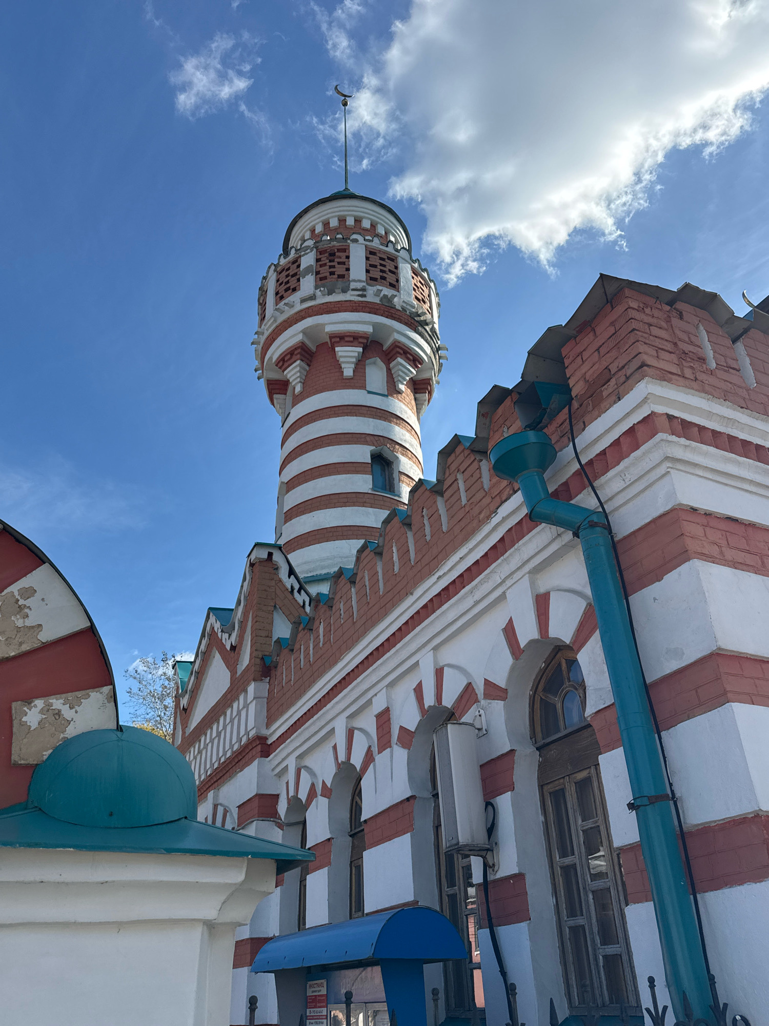 The image shows a low-angle view of the Tver Imperial Traveling Palace in Tver, Russia, under a bright blue sky with scattered clouds. The palace, also known as the Travel Palace, is characterized by its distinctive red and white striped tower and the building's facade, which features alternating red brick and white plaster with red arched accents above the windows. A turquoise downspout is visible running down the side of the building. In the foreground, a section of a white wall with a turquoise domed structure is present on the left. The overall scene depicts a historical architectural landmark on a clear day.