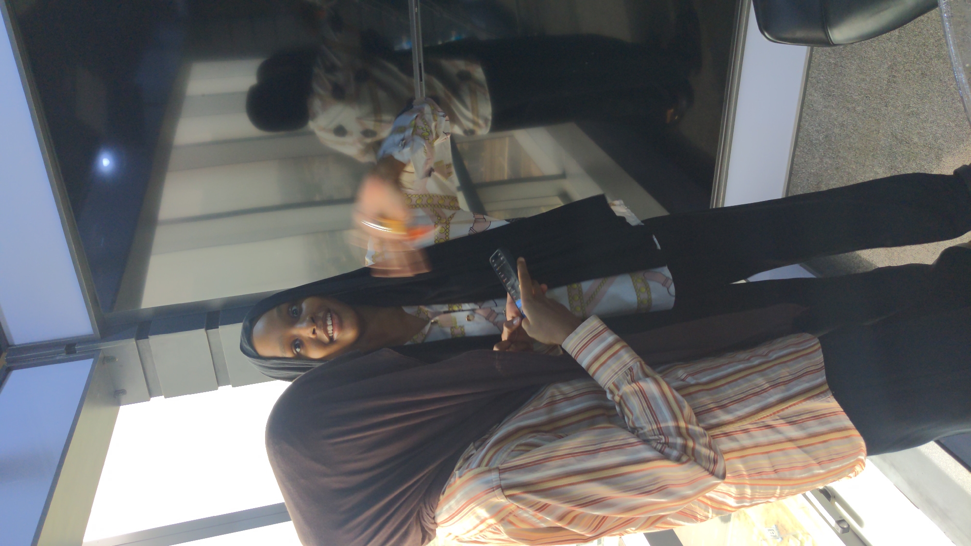 A young woman is captured in a vertical, close-up shot, smiling warmly towards the camera. She wears a dark brown hijab draped over her head and shoulders. Underneath, she has on a light-colored patterned shirt, overlaid with a striped long-sleeved shirt featuring horizontal bands of orange, yellow, white, and brown. Dark pants complete her attire.
In her right hand, she holds a black remote control, pointing it forward and slightly left. Her left hand holds a dark-colored smartphone.
The setting appears to be an indoor space in Abuja, Nigeria, characterized by a large, reflective surface—possibly a window or a mirror—occupying the left side of the frame. This surface reflects a bright light source, suggesting daytime. The reflection also reveals another individual, likely an older woman, standing further back. This reflected person is also dressed in a light-colored patterned shirt and dark pants, and a dark office chair is partially visible in the reflected background. The woman in the foreground conveys a sense of happiness and friendliness through her bright smile. No discernible text is visible in the image.