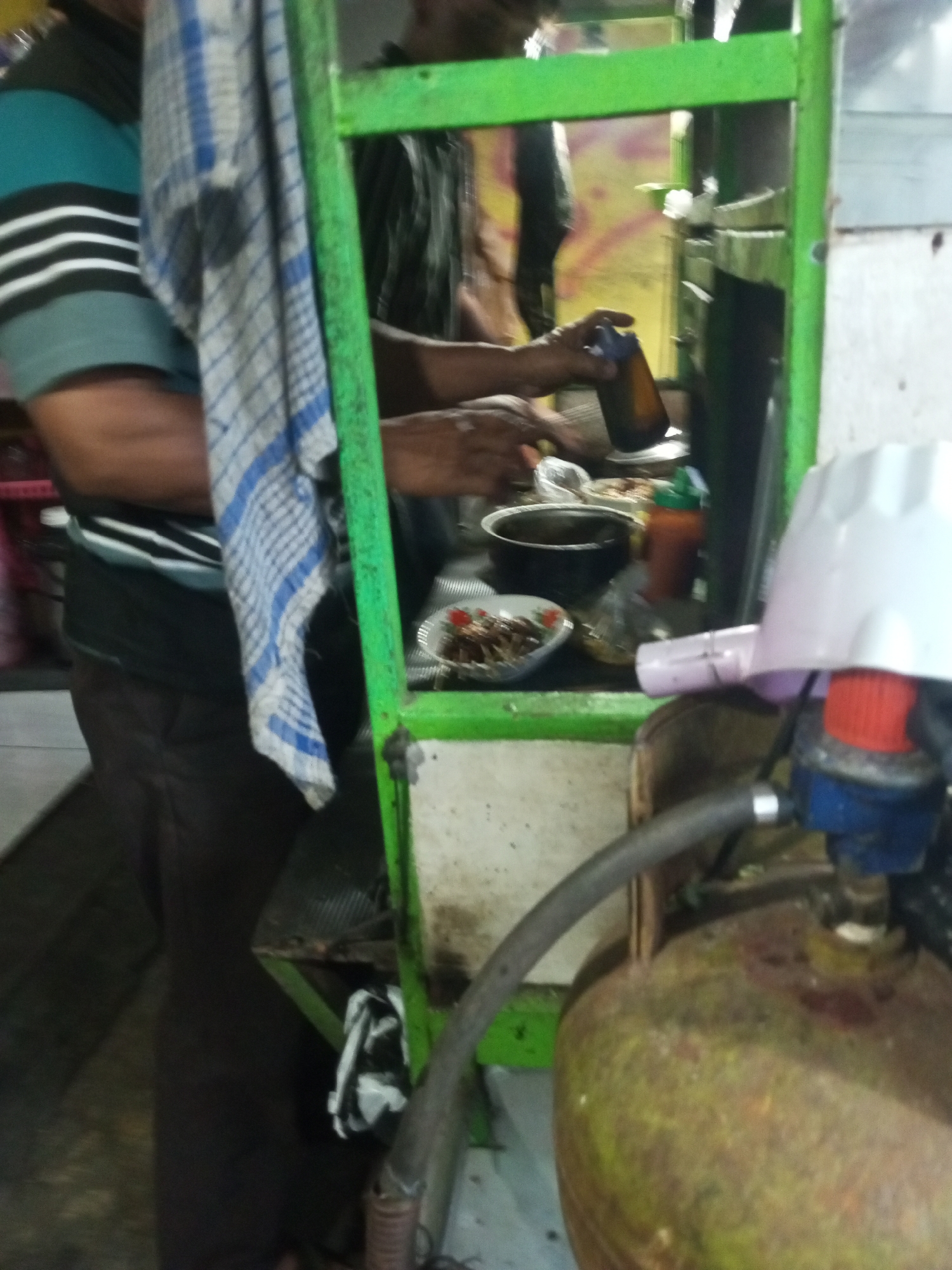 In Andongsili, Indonesia, a food stall is depicted. The stall has a green metal frame. A person wearing a turquoise and black striped shirt is visible on the left. A blue and white checkered cloth hangs from the frame. In the center, a hand holds a brown bottle. A gas tank with a white regulator is in the foreground on the right. Various food items and containers are on display within the stall.