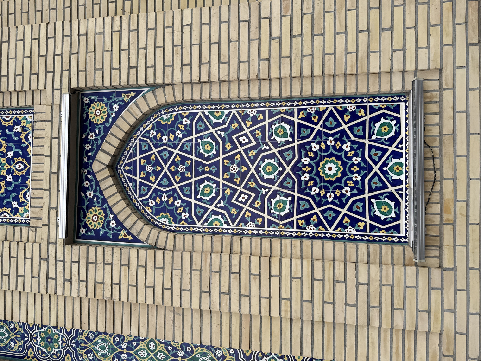 The image shows an arched window with intricate blue, green, and white geometric tilework, set against a light yellow brick wall. The window is framed by a border of similar tilework. A metal ledge runs below the window, with some wires visible. The brickwork is arranged in horizontal courses. The overall style of the tilework suggests an Islamic architectural influence, common in Tashkent, Uzbekistan. The scene is devoid of people and any active events, presenting a static architectural detail.