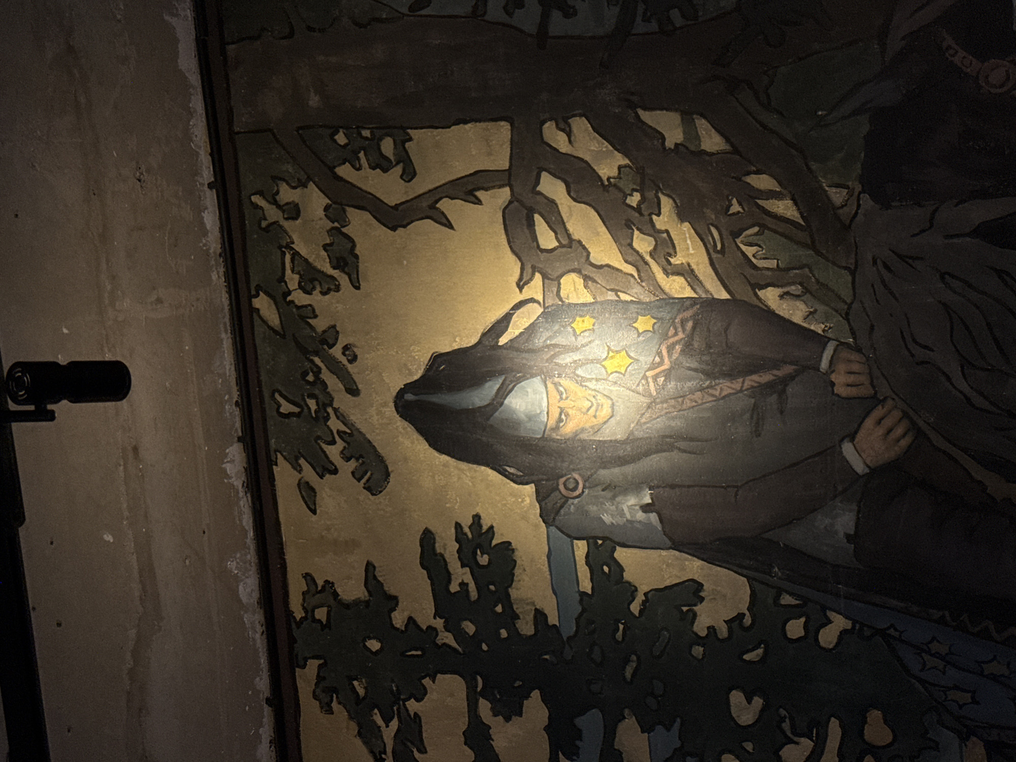 The image shows a mural depicting a man in a pointed blue hat and a dark cloak, with long dark hair falling over his shoulders. He has a determined expression and is illuminated by a spotlight. Two yellow stars are visible on his chest. He appears to be in a forest setting with dark trees. The mural is located on a wall that shows signs of wear and has a spotlight directed at it from above. The mural is part of the interior of the Melikhovo estate in Moscow Oblast, Russia.