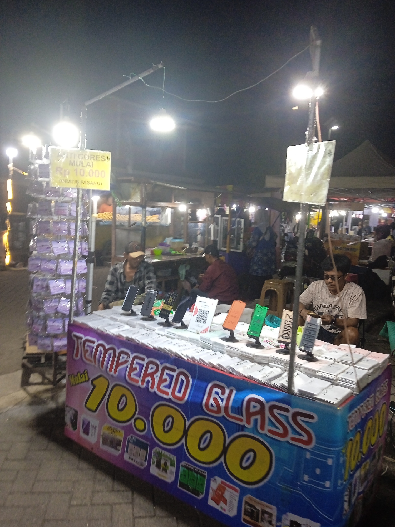 The image shows an outdoor stall at night, likely in Kendal, Indonesia. The stall is selling tempered glass screen protectors for mobile phones. A prominent banner on the stall reads "TEMPERED GLASS MULAI 10.000" (Tempered Glass Starts at 10,000 Rupiah) with additional text indicating "GRATIS PASANG" (Free Installation). Several stacks of tempered glass packages are displayed on the counter. Two men are visible at the stall. One man, wearing a plaid shirt and a cap, appears to be interacting with the display. Another man, wearing a t-shirt and sunglasses, is seated and looking at his phone. In the background, there are other stalls and people, suggesting a bustling night market or street fair. The lighting from the stall's lamps illuminates the area, creating a warm glow against the dark night sky. The ground is paved with interlocking bricks.