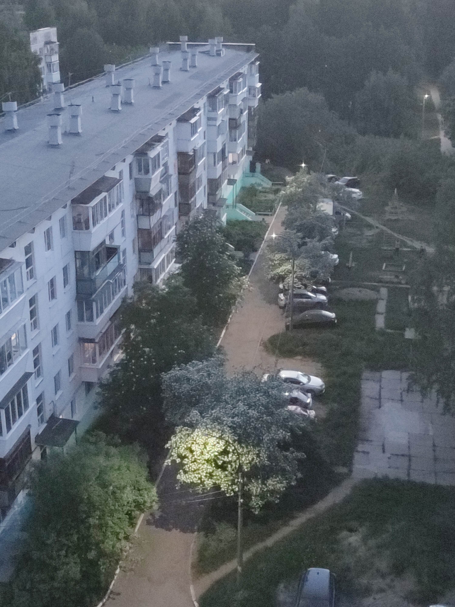 The image depicts an elevated view of a residential area. A long, multi-story apartment building with a gray roof and white facade dominates the left side of the frame. The building features numerous windows and balconies. A paved walkway runs parallel to the building, lined with trees. Several parked cars are visible alongside the walkway, interspersed with trees and grassy areas. The lighting suggests it is either dusk or dawn. The setting appears to be a city, indicated by the residential buildings and infrastructure. The location is Unknown, Unknown. No people are discernible in the image.