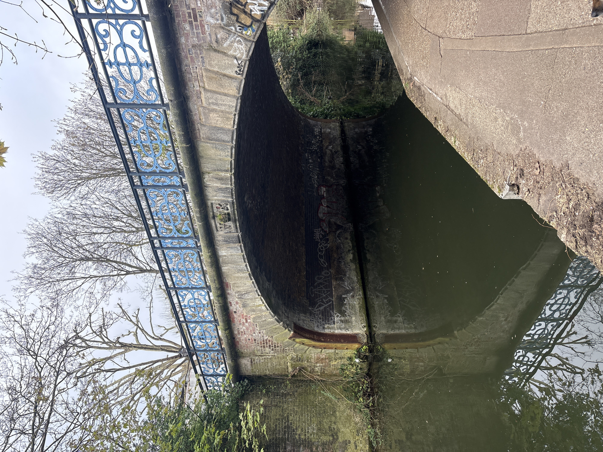 The image depicts a canal bridge in London, United Kingdom. The bridge is constructed of brick and concrete, with a blue ornate metal railing on one side. Graffiti is visible on the underside of the arch. The canal water is dark and reflects the bridge and surrounding vegetation. Trees are visible on either side of the canal, some with bare branches. The sky is overcast. There are no people visible in the image.