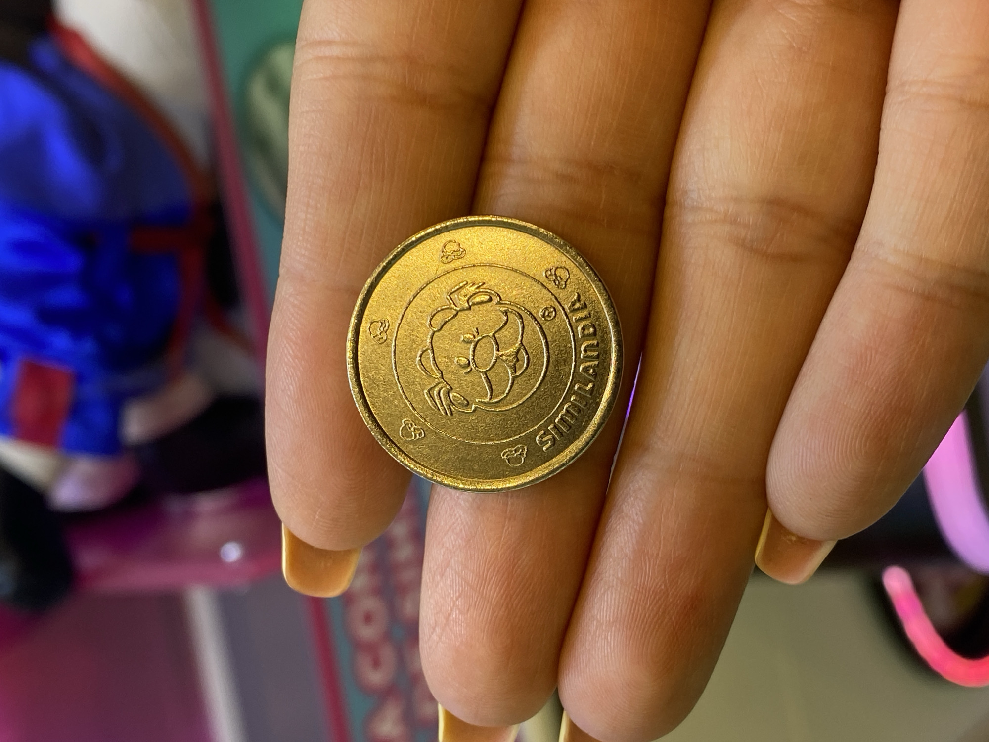 A close-up shot features a gold-colored coin resting on the fingertips of a human hand. The coin appears to have a cartoon animal design on its face with text around the perimeter that includes the words "Similandia". The hand's fingernails are painted with a light-colored manicure. The background contains indistinct elements of a toy or arcade game. The location within the city of San Bernardino Tlaxcalancingo, Mexico, cannot be definitively determined from the image content alone.