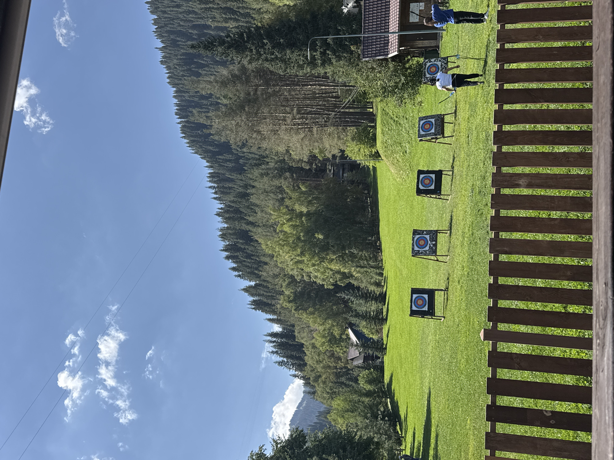 An outdoor archery range is set up on a grassy hill in a mountainous area, likely in or near Almaty, Kazakhstan. The scene captures a sunny day with a clear blue sky dotted with a few white clouds. In the foreground, a wooden fence runs horizontally across the frame, obscuring the lower portion of the image. Beyond the fence, a lush green lawn stretches towards a line of archery targets. There are four targets visible, each with concentric circles of red, blue, and white.
In the mid-ground, two individuals are engaged in archery. One person, wearing a white shirt and dark pants, is standing with a bow and arrow, facing away from the camera and towards a target. Another person, dressed in a blue shirt and dark pants, is also holding a bow and arrow, appearing to be in the process of shooting. Behind the archery targets, the landscape rises into a dense forest of evergreen and deciduous trees, with a few scattered buildings nestled amongst them, suggesting a resort or recreational area. A dark-colored building with a brown roof is visible to the right, and further back, a larger wooden house is partially obscured by trees. The background is dominated by the steep, forested slopes of a mountain range, indicative of the natural beauty surrounding Almaty. Power lines are faintly visible stretching across the upper portion of the sky, and a lamppost stands near the building on the right. The overall impression is one of a serene, nature-filled recreational setting.