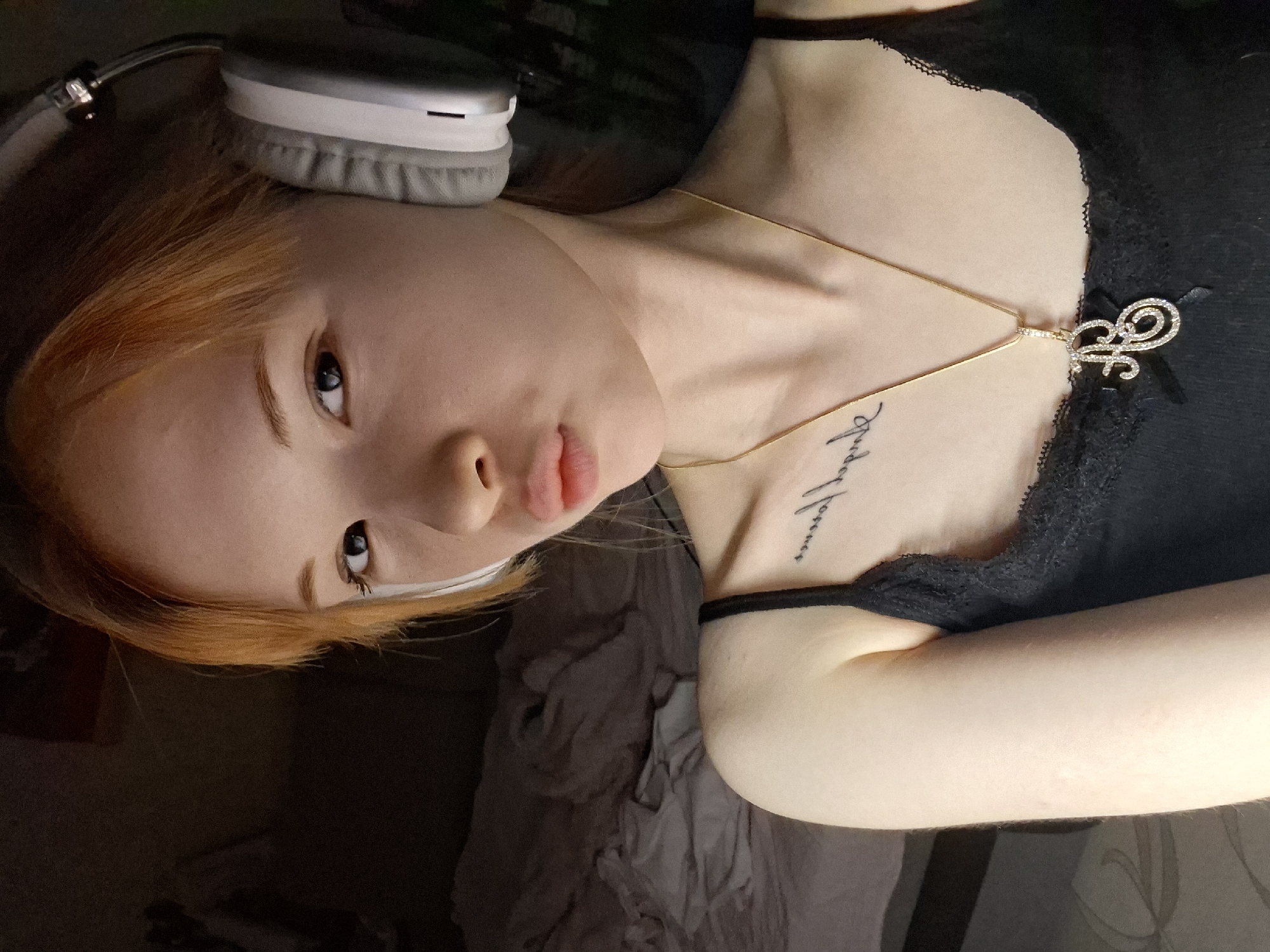 A young person, likely female, with fair skin and light reddish-blonde hair, is depicted in a close-up shot, rotated 90 degrees clockwise as if lying down or leaning back. She has blue eyes and gazes directly at the camera with a slightly pouting expression. A white strip or tape is visible near her left eye.
She wears light grey and white over-ear headphones. Around her neck is a delicate golden chain necklace, featuring a sparkling, ornate pendant that appears to be a stylized initial, possibly an 'H' or 'J', embellished with rhinestones and a small black bow. She is dressed in a black top with a lace-trimmed neckline, and a dark strap (possibly from the top or a separate accessory) is visible on her left shoulder. On her right collarbone/upper chest area, there is a tattoo written vertically in a script font, which clearly spells out "Forever."
The background is dark and blurry, suggesting an indoor setting with subdued lighting, possibly evening or night. Indistinct shapes in the background hint at bedding or furniture. The image captures a quiet, intimate moment, possibly of the subject listening to audio. There are no specific visual cues within the image that denote its location as Chaikovsky, Russia.