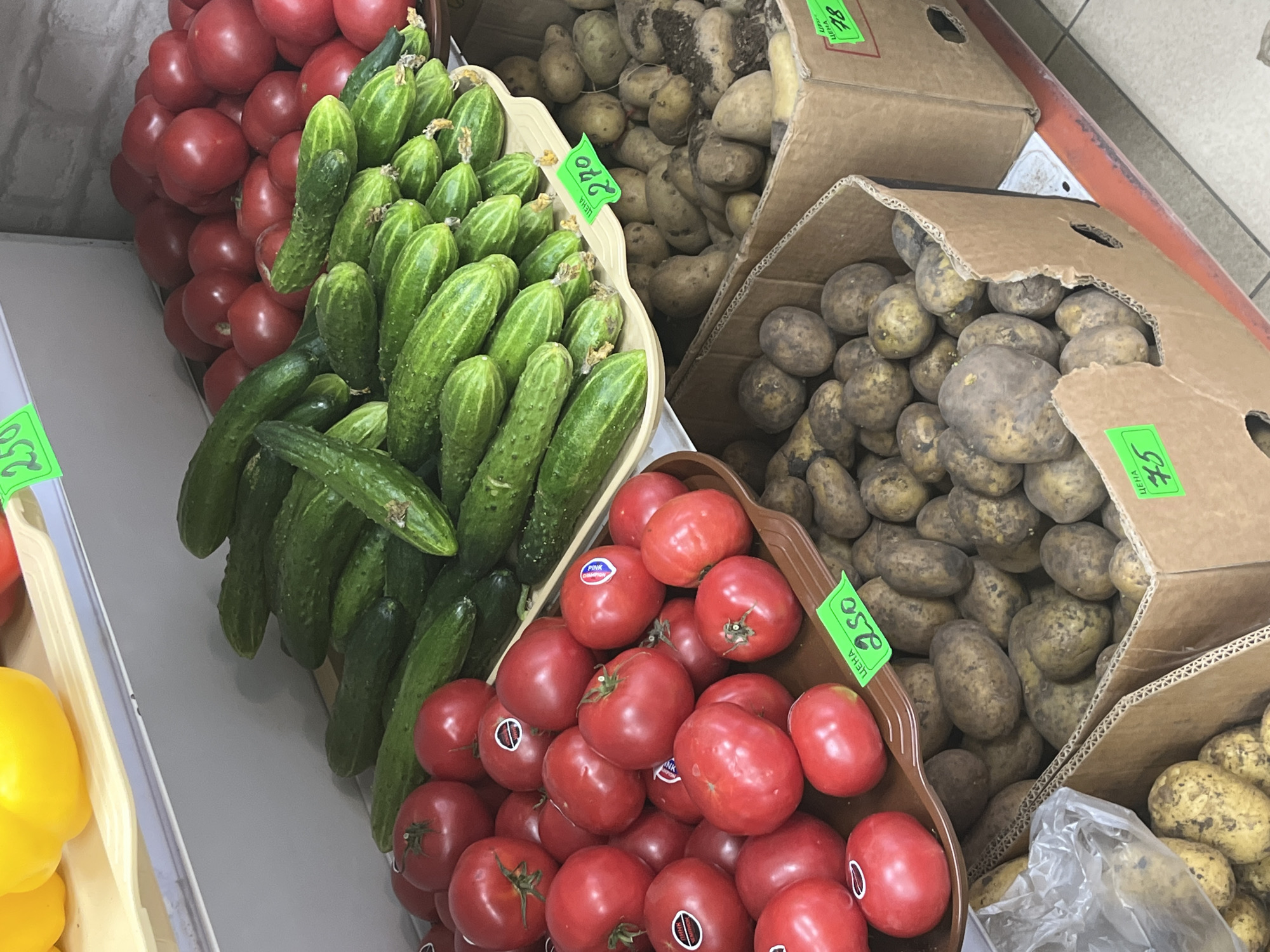 This image depicts a produce display at a market in Sochi, Russia. In the foreground, several cardboard boxes are filled with potatoes. The box on the left contains potatoes with a lighter skin, while the box on the right holds potatoes with a darker, more earthy appearance. A plastic bag filled with potatoes is also visible to the left of the boxes. A brown tray, positioned above the potatoes, holds a large quantity of red tomatoes. These tomatoes are round and appear ripe, with some having white stickers indicating they are a specific variety. To the left of the tomatoes, a bright yellow bell pepper is partially visible on a white shelf. Behind the tomatoes, a large pile of green cucumbers is neatly arranged on a cream-colored tray. Several price tags are visible throughout the display. One green tag with black text reading "250" is attached to the tray of tomatoes. Another green tag, also with black text, reads "270" and is placed near the cucumbers. A third green tag reads "75" and is attached to the box of darker potatoes. The background shows a white brick wall. The overall scene suggests a bustling market environment with fresh produce for sale.
