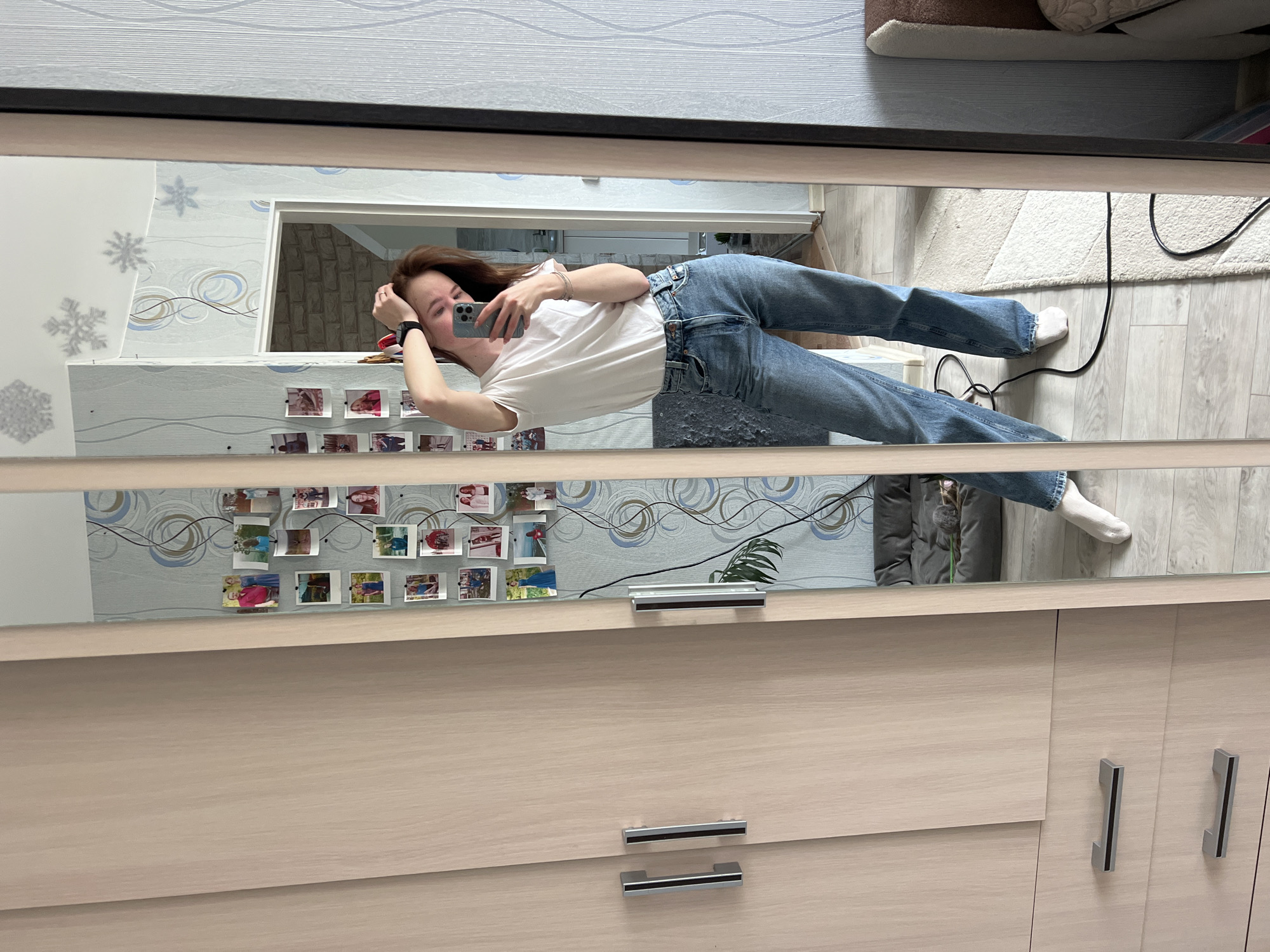 A young woman is captured in a mirror selfie, standing or leaning in an indoor setting. She has long, dark hair, and is wearing a white t-shirt, blue jeans, and white socks. She holds a gray smartphone in her right hand, taking the picture, and wears a black smartwatch on her left wrist. Her expression appears neutral, with a slight hint of a smile.
The mirror reflects a room adorned with light-colored wallpaper featuring grey snowflakes and abstract blue and brown swirling patterns. Numerous small, square-shaped printed photographs are casually taped to the wallpaper, showcasing various people and scenes. A window or opening is visible in the reflected background, along with a partially seen potted plant. The floor is covered with light wood-patterned laminate or hardwood, on which a white shaggy rug and some black electrical cables are visible. Part of a brown, upholstered object, possibly a pet bed or another piece of furniture, is also reflected. The mirror itself is set into a light wood-grain wardrobe or cabinet, equipped with silver handles.
There is no discernible text visible in the image. The time of day and external weather conditions cannot be determined from the indoor lighting. The scene suggests a personal residential space, likely in Chaikovsky, Russia.