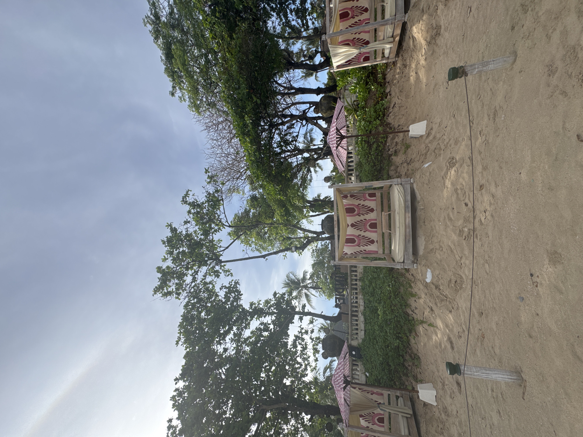The image depicts a beach scene in Kuta Selatan, Indonesia. Several wooden beach cabanas with decorative fabric roofs are lined up along a sandy area with sparse vegetation. The scene is partially shaded by various trees, including a palm tree. A pink umbrella is visible between two cabanas. Wooden poles with ropes extend along the beach, likely marking boundaries or walkways. The sky is light blue with thin clouds. No people are visible.
