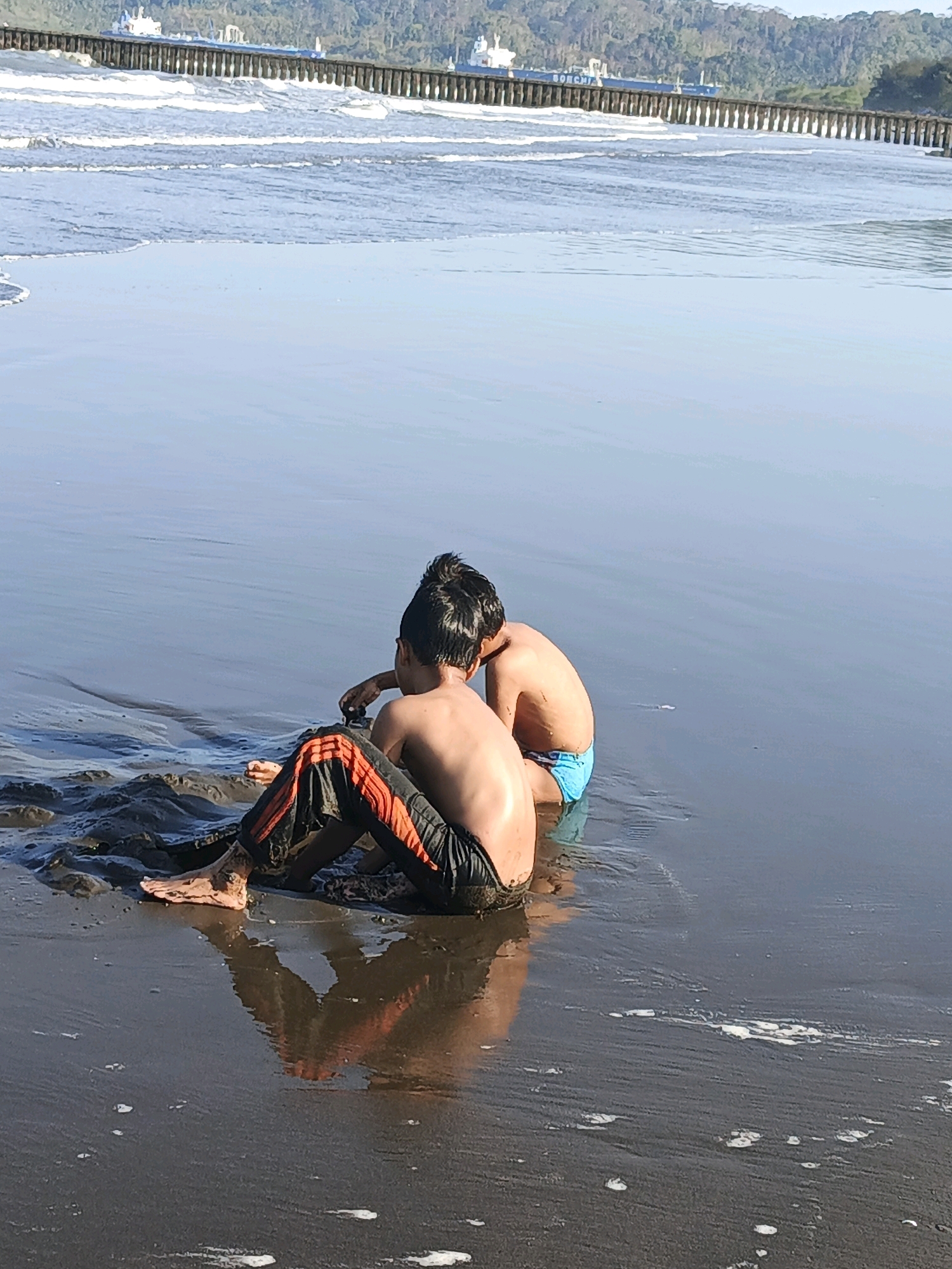 Two young boys are sitting on a dark, wet sand beach, partially in the shallow water. The boy on the left is wearing black pants with orange stripes. The boy on the right is wearing blue swim trunks. The water is calm with gentle ripples. In the background, there is a wooden pier extending into the sea, and beyond that, a line of ships and a tree-covered hill under a bright sky. The reflection of the boys and the pier are visible in the wet sand. The scene appears to be in Kelurahan Cilacap, Kecamatan Cilacap, Indonesia.