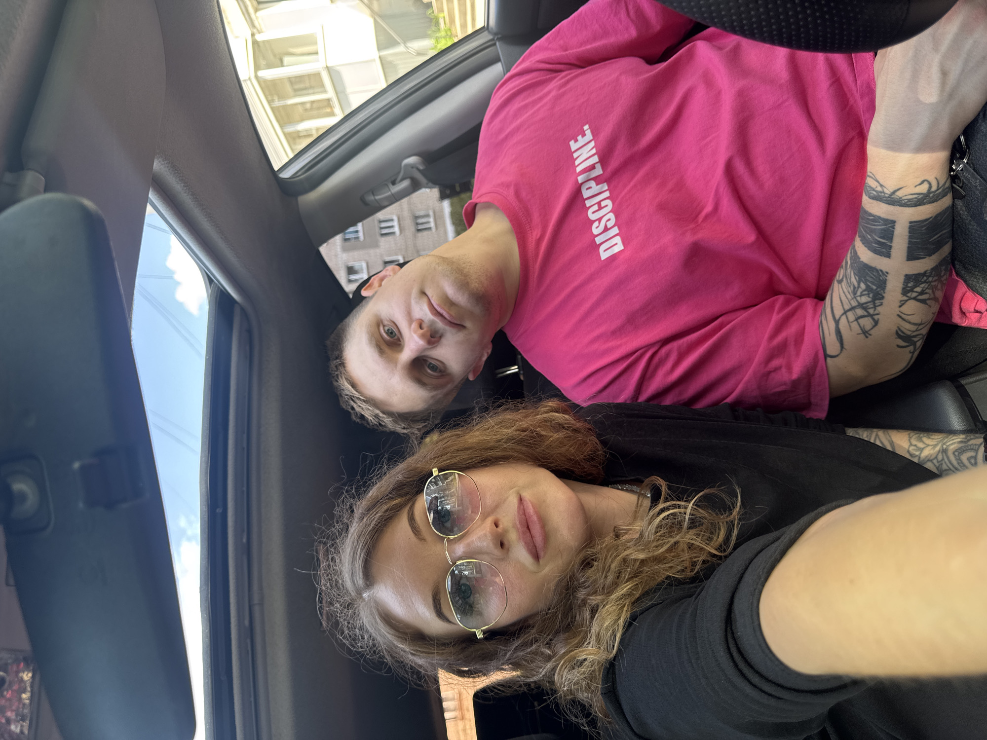 Two individuals are captured in a selfie inside a car, potentially in the city of Chaikovsky, Russia, considering the potential location information from the prompt context. The man, positioned towards the upper portion of the frame, is wearing a bright pink long-sleeved shirt with the word "DISCIPLINE" printed on it. He has fair skin, light hair, and is looking into the camera. His left forearm is visible, featuring several black tattoos. The woman, located below him, has wavy brown hair and is wearing gold-rimmed sunglasses. She is dressed in a black top. The car features a sunroof, through which glimpses of buildings and the sky are visible, suggesting an urban setting. The interior of the car is predominantly dark.