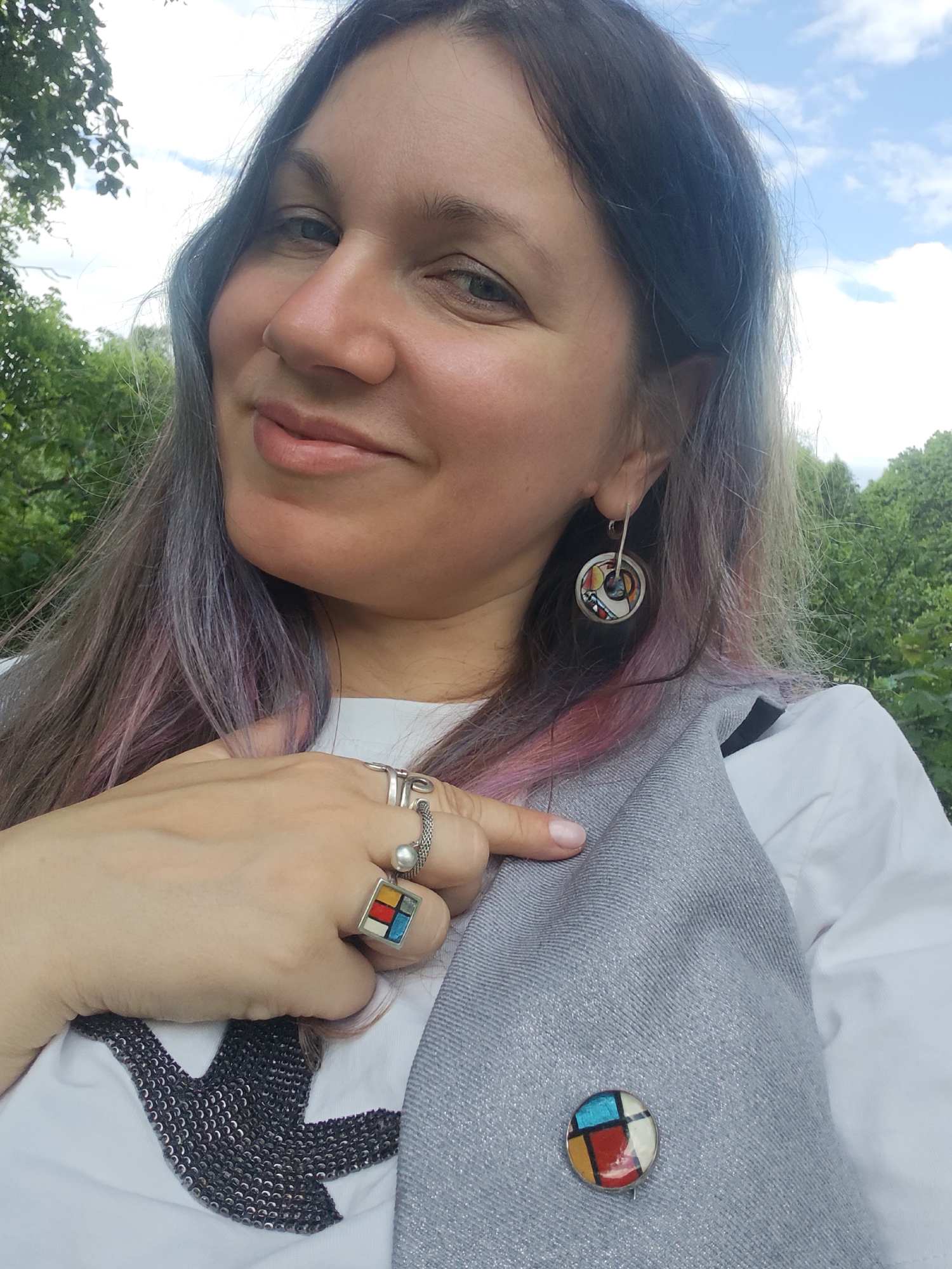 A woman is posing outdoors. She has long hair with some sections dyed purple and pink, and is wearing earrings and multiple rings. She is pointing with her right hand towards a pin on her grey vest. She is also wearing a white shirt with a sequined design. In the background, there are green trees and a partly cloudy sky. There is no verifiable information to suggest this image was taken in Selivanovo, Russia.