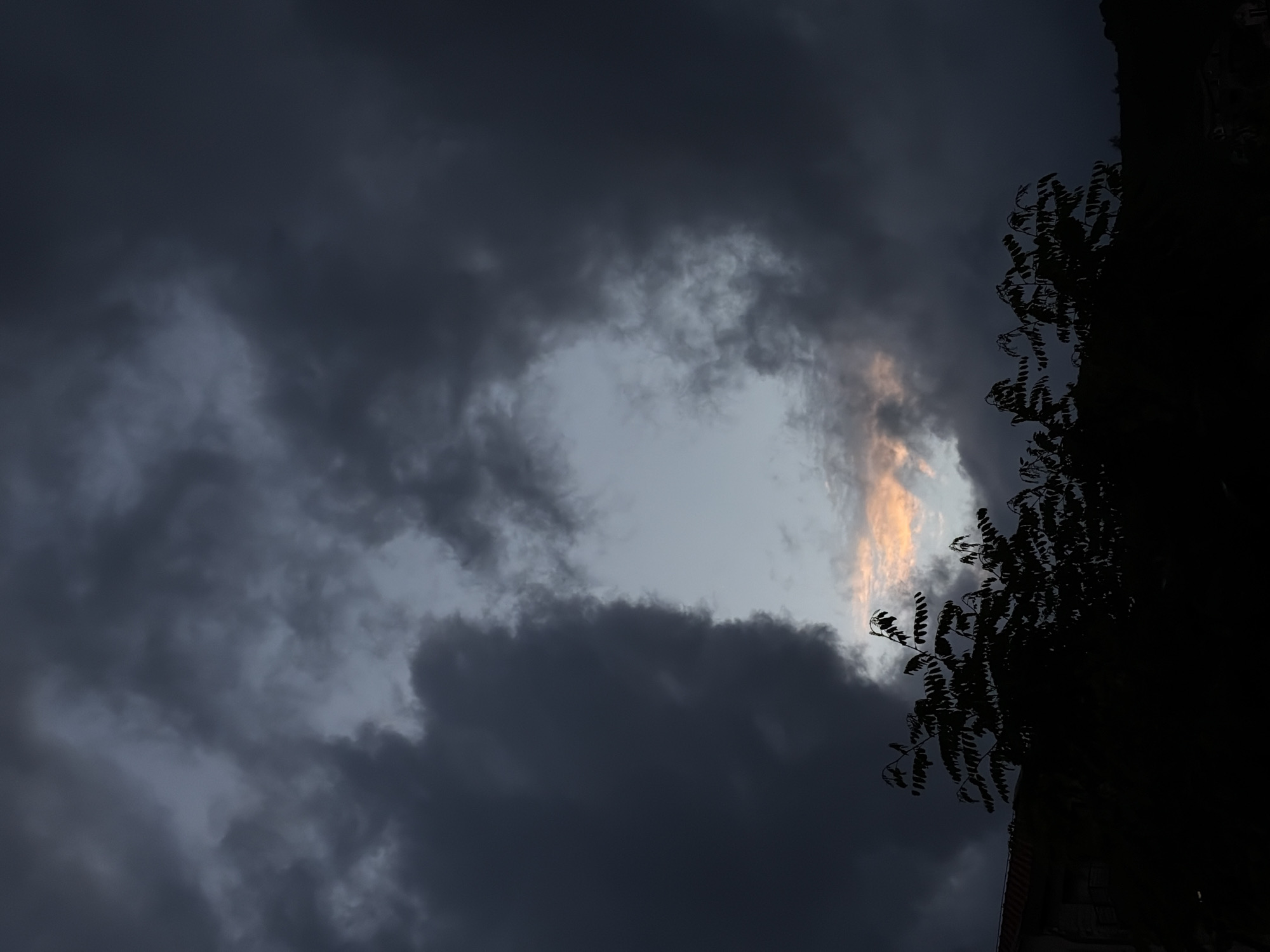 The image shows a view of the sky with dark, stormy clouds, with a small patch of lighter sky visible through an opening in the clouds. A tree silhouette is visible on the right side of the frame, along with part of a building. The scene appears to be taken in Budva, Montenegro.