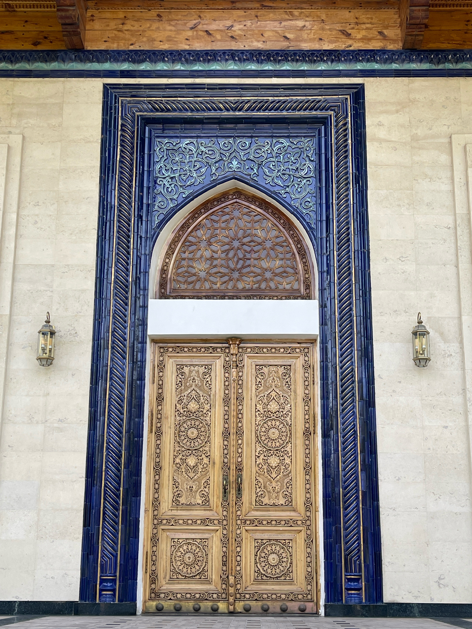 The image shows the grand entrance of a building in Tashkent, Uzbekistan, likely a mosque or a government building due to its ornate architecture. The central focus is a pair of large, intricately carved wooden doors, golden in color, with detailed floral and geometric patterns. Above the doors is a semicircular arched window with a complex wooden lattice work, also featuring geometric star patterns. The entire entrance is framed by a deeply recessed architectural surround. The inner frame is adorned with vibrant blue ceramic tiles, featuring raised, swirling arabesque patterns in a lighter blue and gold. The outer frame is a darker, textured blue with gold accents. The surrounding walls are clad in large, light-colored stone tiles. On either side of the entrance, mounted on the wall, are traditional lantern-style lights with glass panels and metal frames. The ceiling above the entrance is composed of exposed wooden beams. The ground in front of the entrance is paved with geometric patterned tiles. The image is taken from a frontal, slightly low angle, emphasizing the scale and grandeur of the entryway.