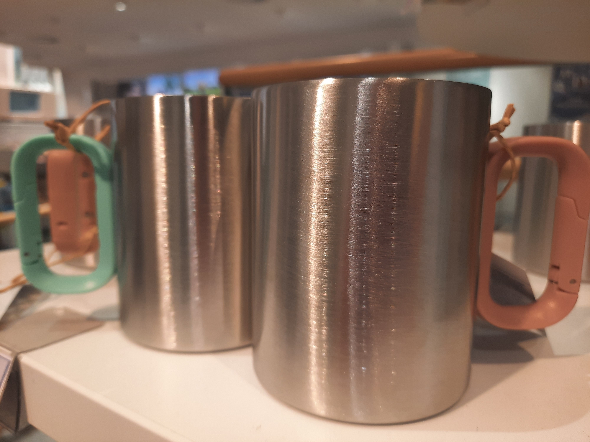 Two stainless steel mugs with colorful carabiner-style handles are displayed on a white surface, likely a shelf in a retail environment. The mug on the right has a pink handle, and the mug on the left has a teal handle. Both handles are attached to the mugs with a thin, tan cord. The mugs have a brushed metal finish and are slightly reflective. The background is blurred, showing shelves and possibly other merchandise, suggesting an indoor commercial setting. The lighting in the scene is bright, causing reflections on the metal surfaces of the mugs. The image appears to have been taken in a store. No individuals are visible in the image. The location is not specified in the image or provided context.