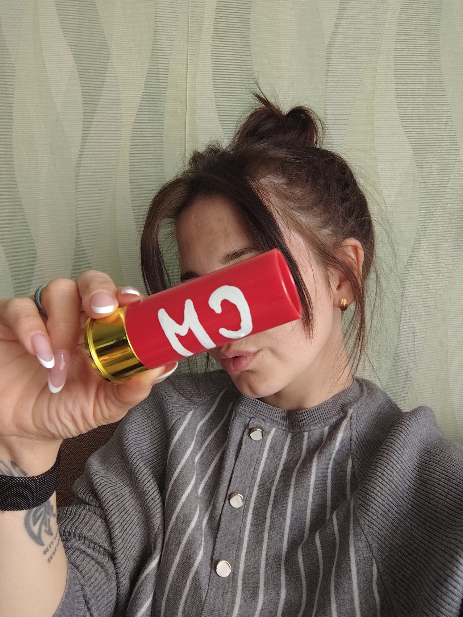 A woman with brown hair in a messy bun is holding a red and gold tube with white letters in front of her face. She is wearing a gray and white striped sweater and has French manicured nails. A tattoo is visible on her left arm. The background is a light green wall with a subtle striped pattern. The location is Фоки, Russia.